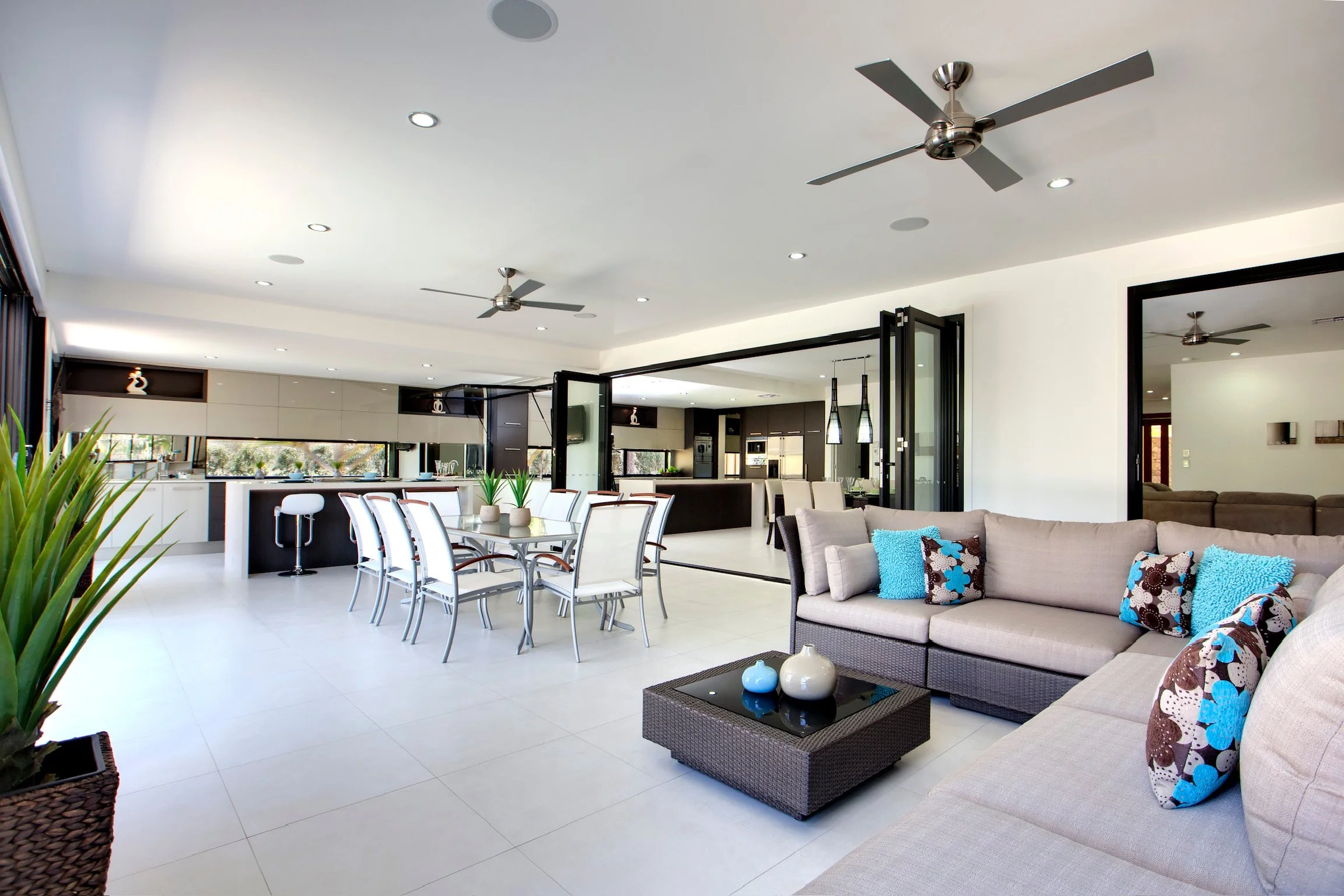 Modern open-concept living and dining area with white walls, a white ceiling with recessed lighting, ceiling fans, a dining table with white chairs, a kitchen with dark cabinets, and a sitting area with beige couches and colorful pillows.