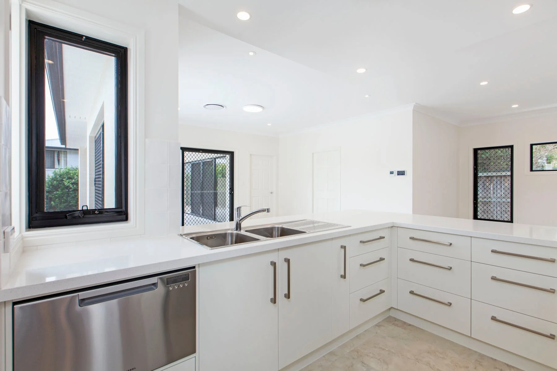 Modern kitchen with white cabinets, stainless steel dishwasher, and black-framed windows, featuring a double sink and a white countertop.