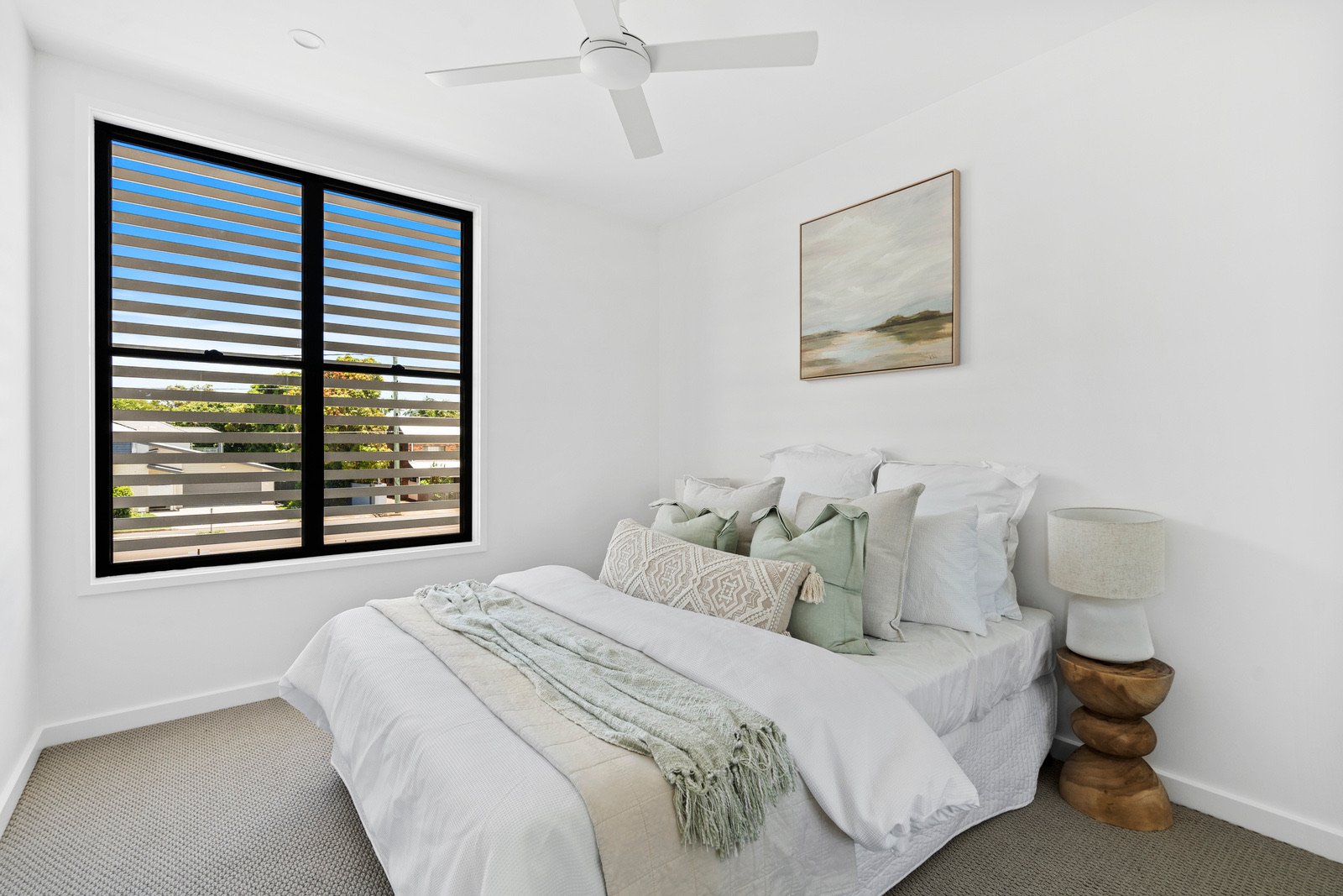 A bedroom with a bed covered in white linens and decorated with multiple pillows, a wooden side table with a lamp, a large window with striped blinds, and a framed landscape painting on the wall.