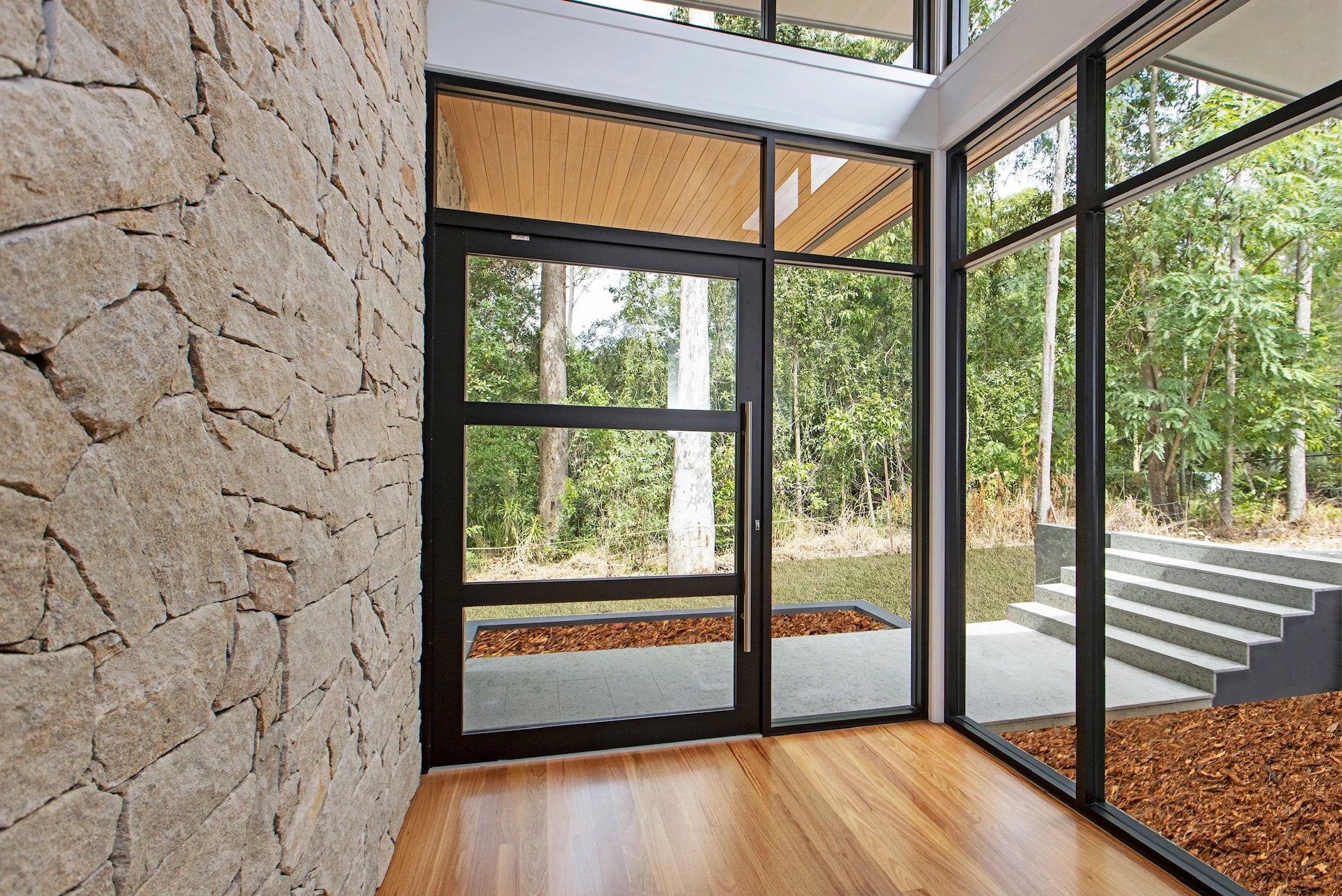 Interior view of a modern house with large glass windows and doors, a stone wall on the left, wooden flooring, and a view of a forest outdoors.