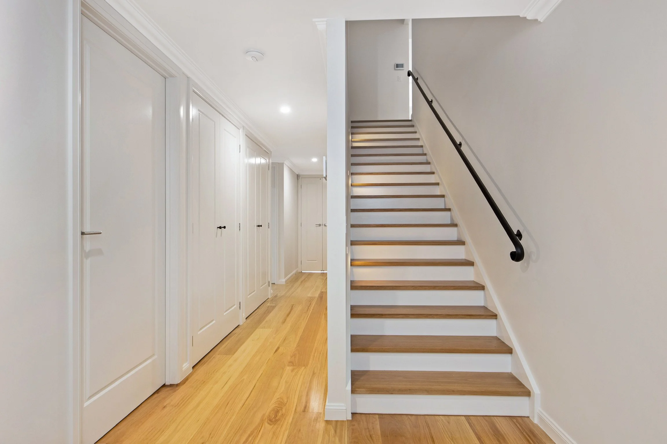 Interior view of a staircase with wooden steps and a black handrail, next to a hallway with white paneled doors and light-colored hardwood flooring.