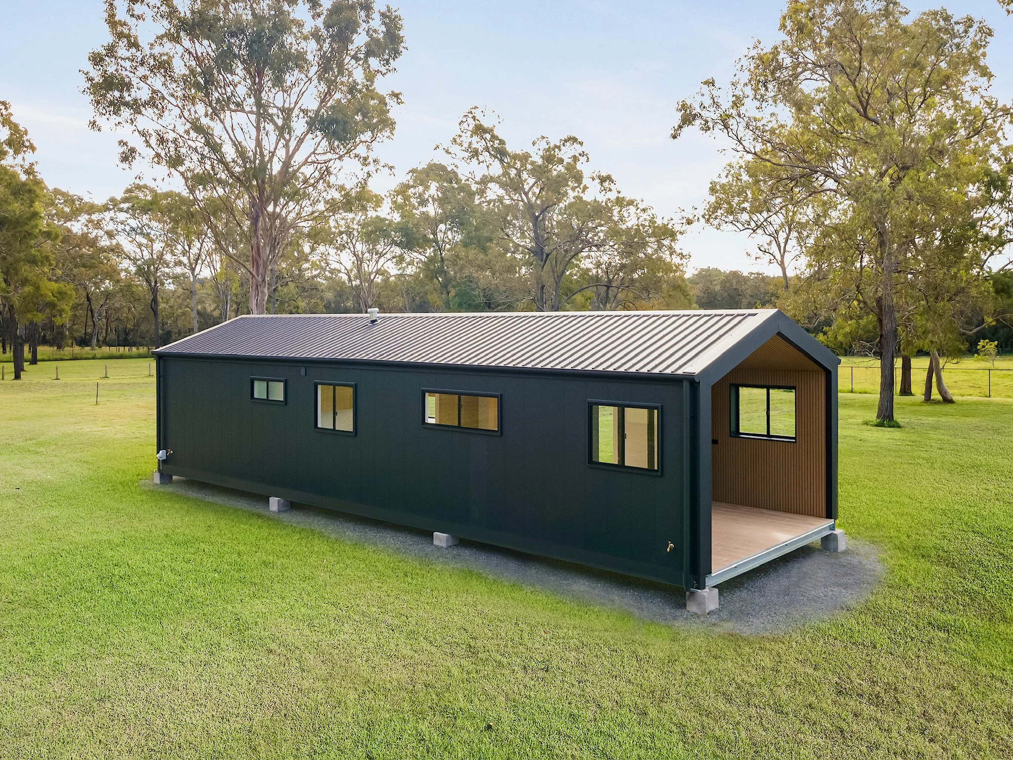 A modern, dark-colored tiny house on stilts with large windows, situated on a lush green lawn surrounded by tall trees under a partly cloudy sky.