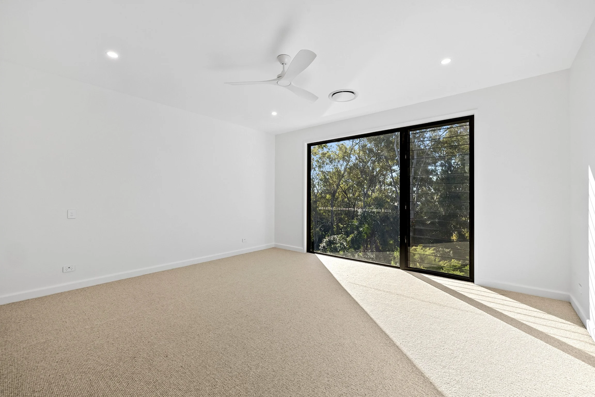 Empty room with beige carpet, white walls, ceiling fan, and large glass sliding door showing trees outside.