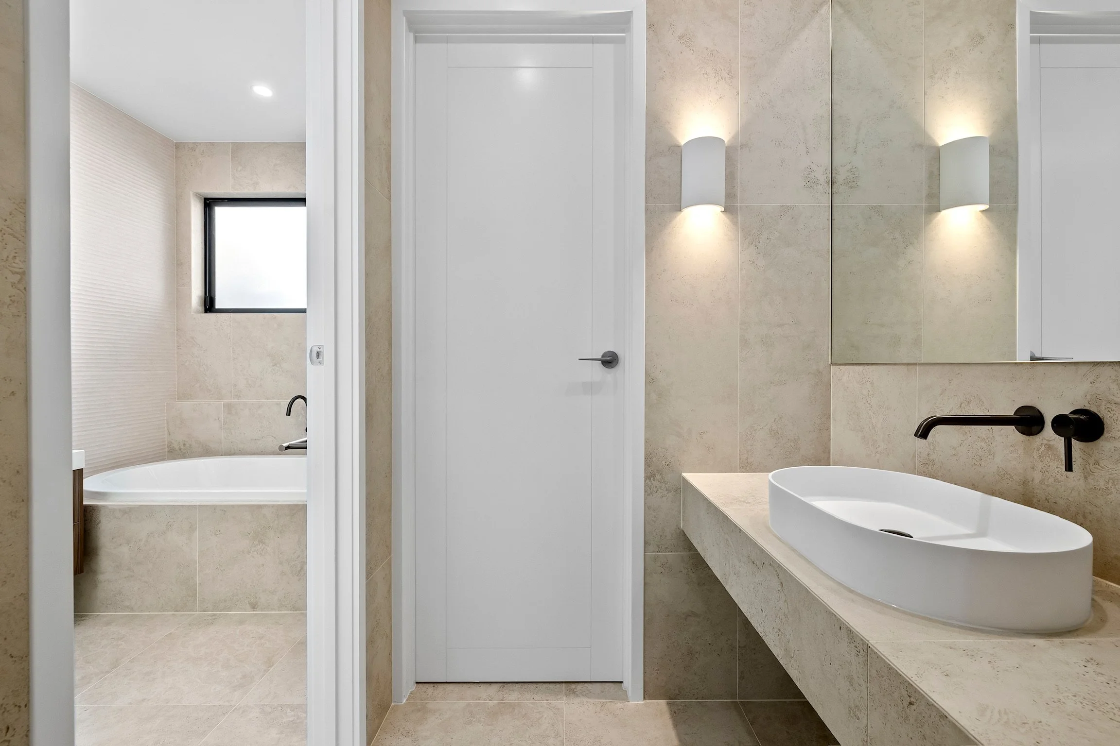 Modern bathroom with beige tiles, a white door, and black fixtures. Features a black-framed window, wall-mounted lights, and a freestanding white bathtub.