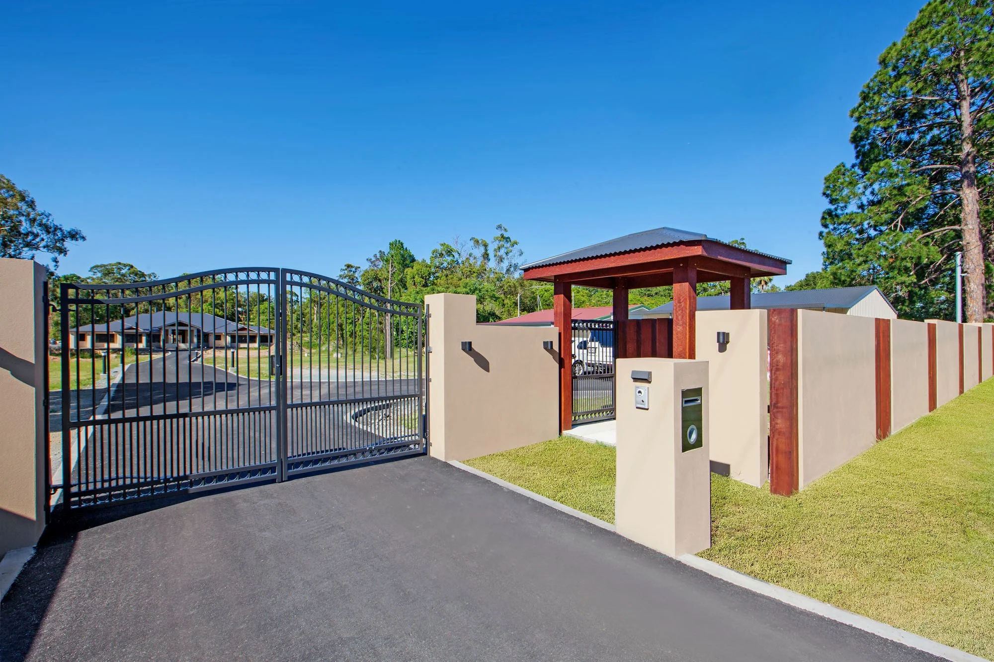 View of a gated entrance to a residential community with a security booth, sidewalk, and a brown wooden fence, surrounded by tall trees and houses in the background under a clear blue sky.