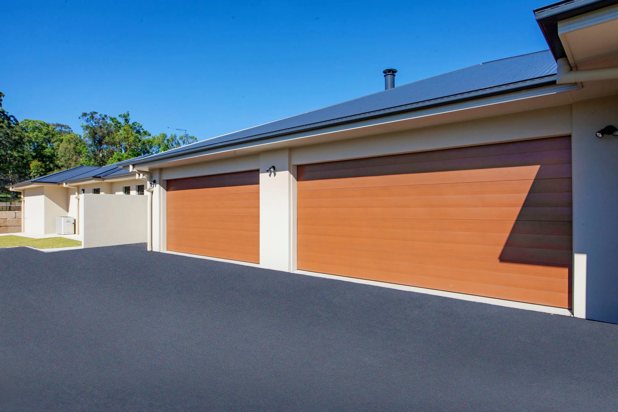 Photo of a row of houses with brown garage doors, white walls, black asphalt driveway, exterior lights, and a blue sky with trees in the background.