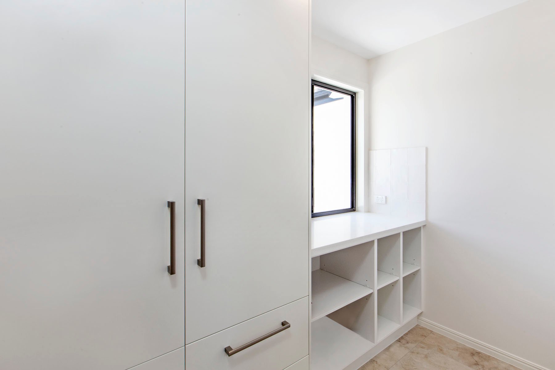 A minimalistic white cabinet with silver handles next to a small window with a black frame, opening to the outside, in a bright room with white walls and beige tiled flooring.