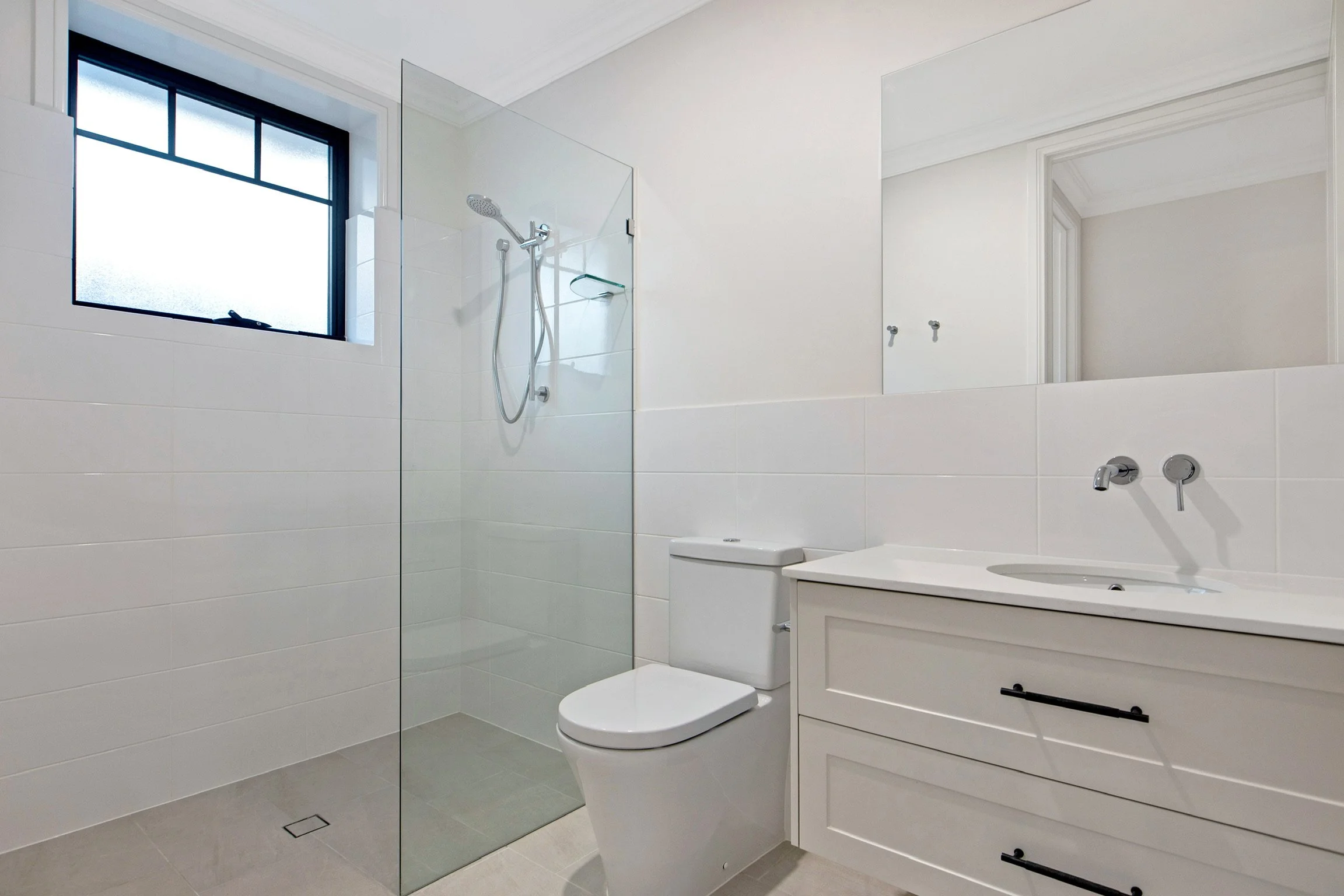 Modern bathroom with a frosted window, shower area with glass partition, white toilet, and white vanity cabinet with black handles.