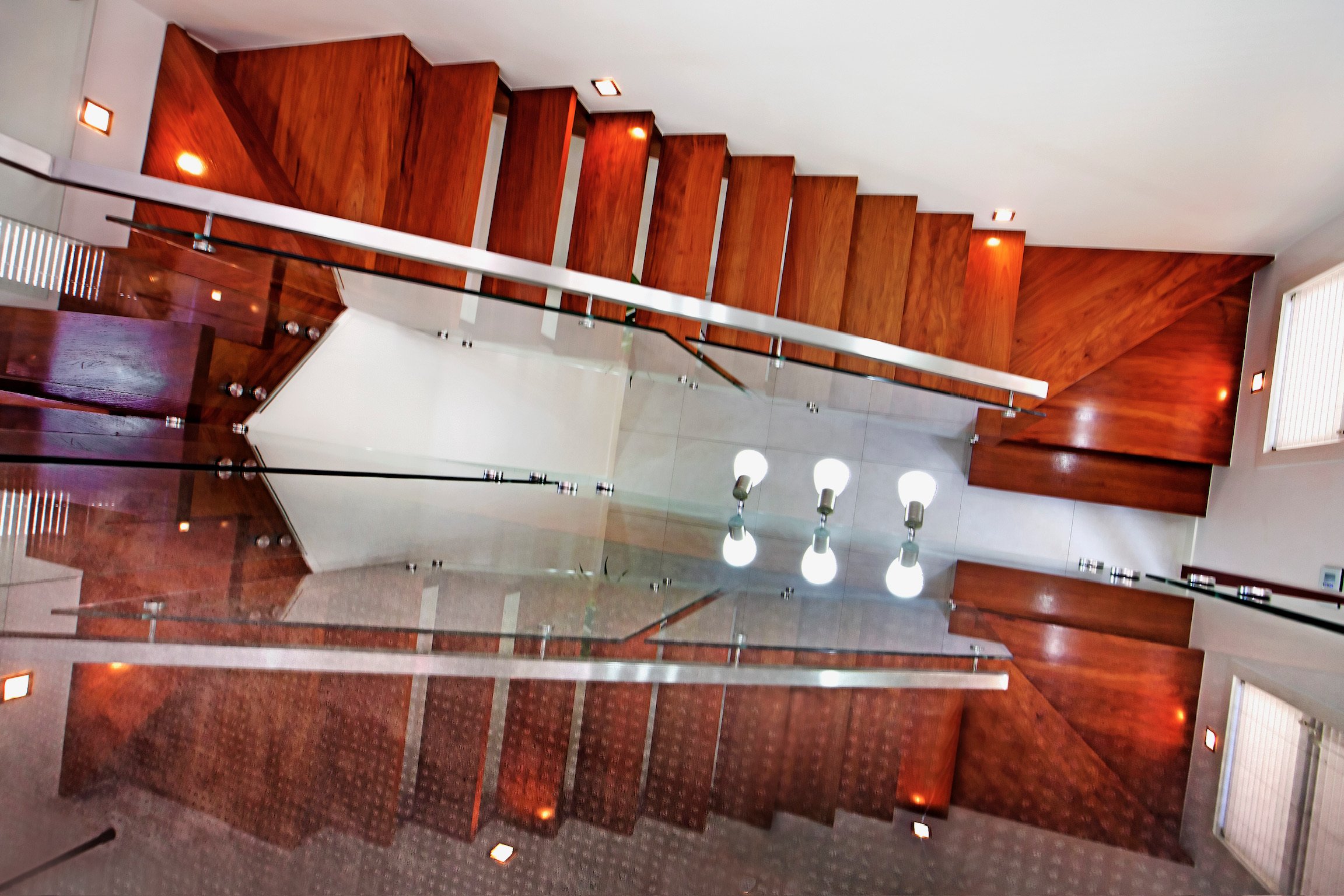 An indoor staircase made of polished wood with a glass and metal railing, leading to an upper floor with a white ceiling and recessed lighting. The lower area features a red carpeted floor and white tiled section.