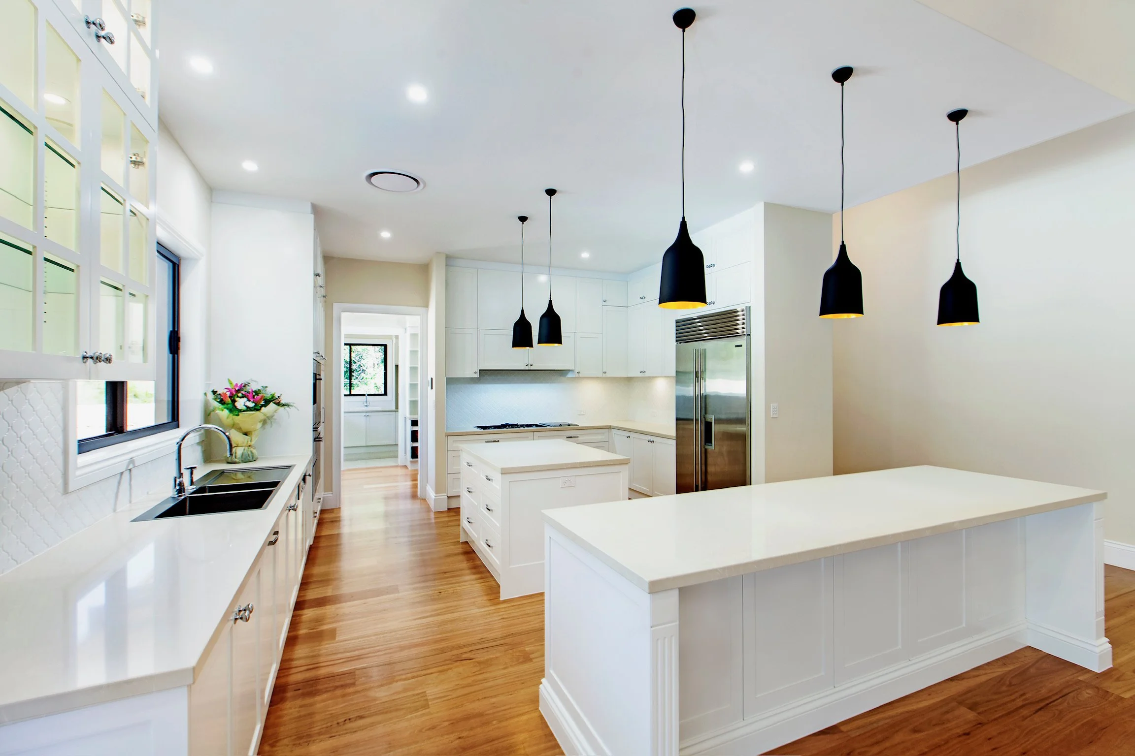 Bright modern kitchen with white cabinets and countertops, black pendant lights hanging over a kitchen island, hardwood floors, and large windows allowing natural light.