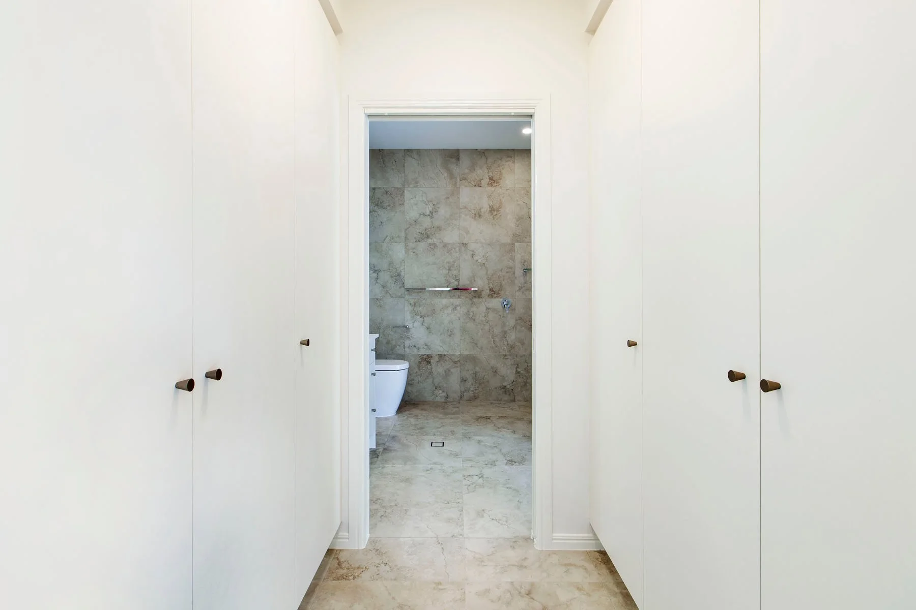 View through a closet door opening to a bathroom with beige marble tiles, a white toilet, and a wall-mounted grab bar.