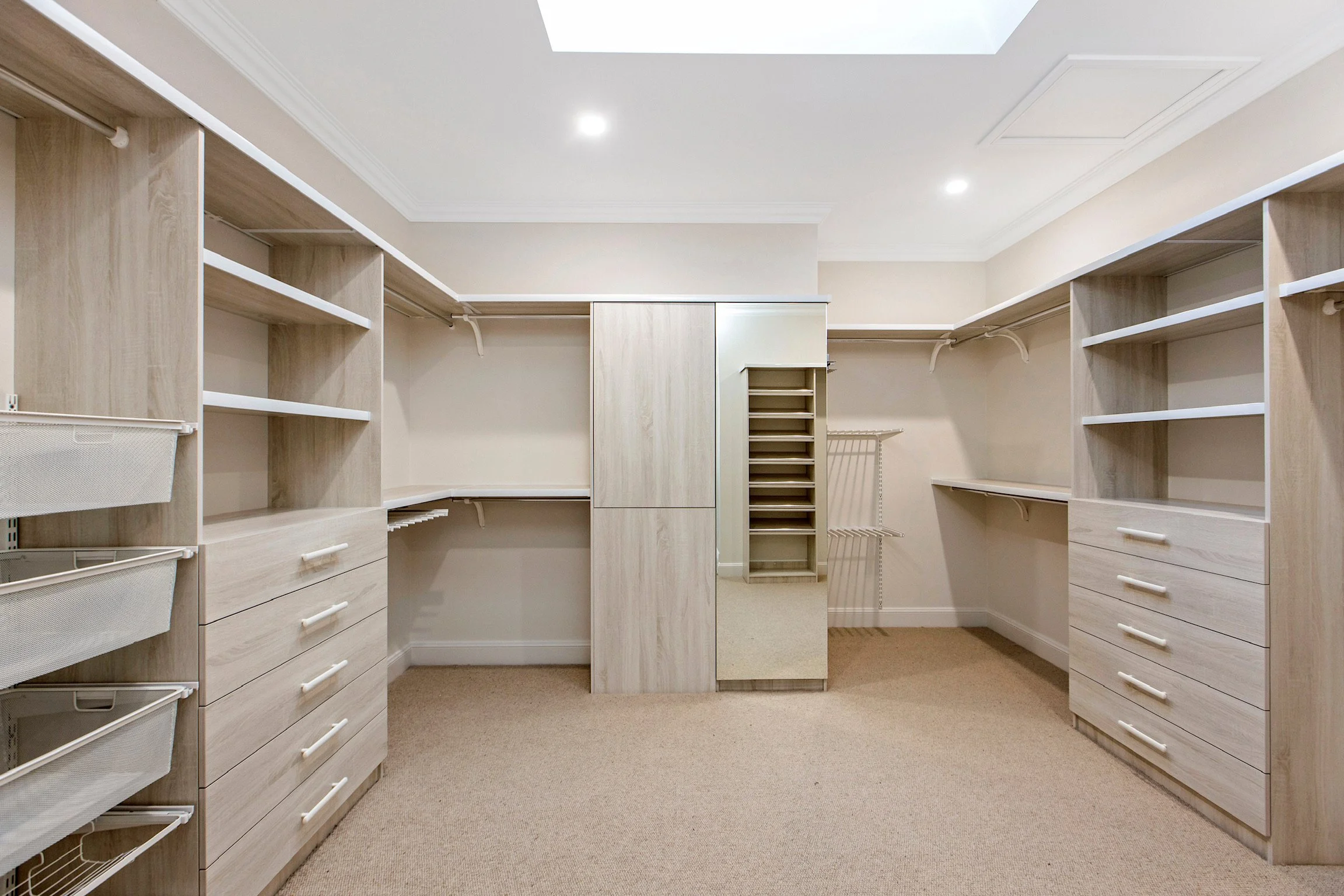 Empty walk-in closet with white shelves and drawers on both sides, beige walls, carpeted floor, and a skylight on ceiling.