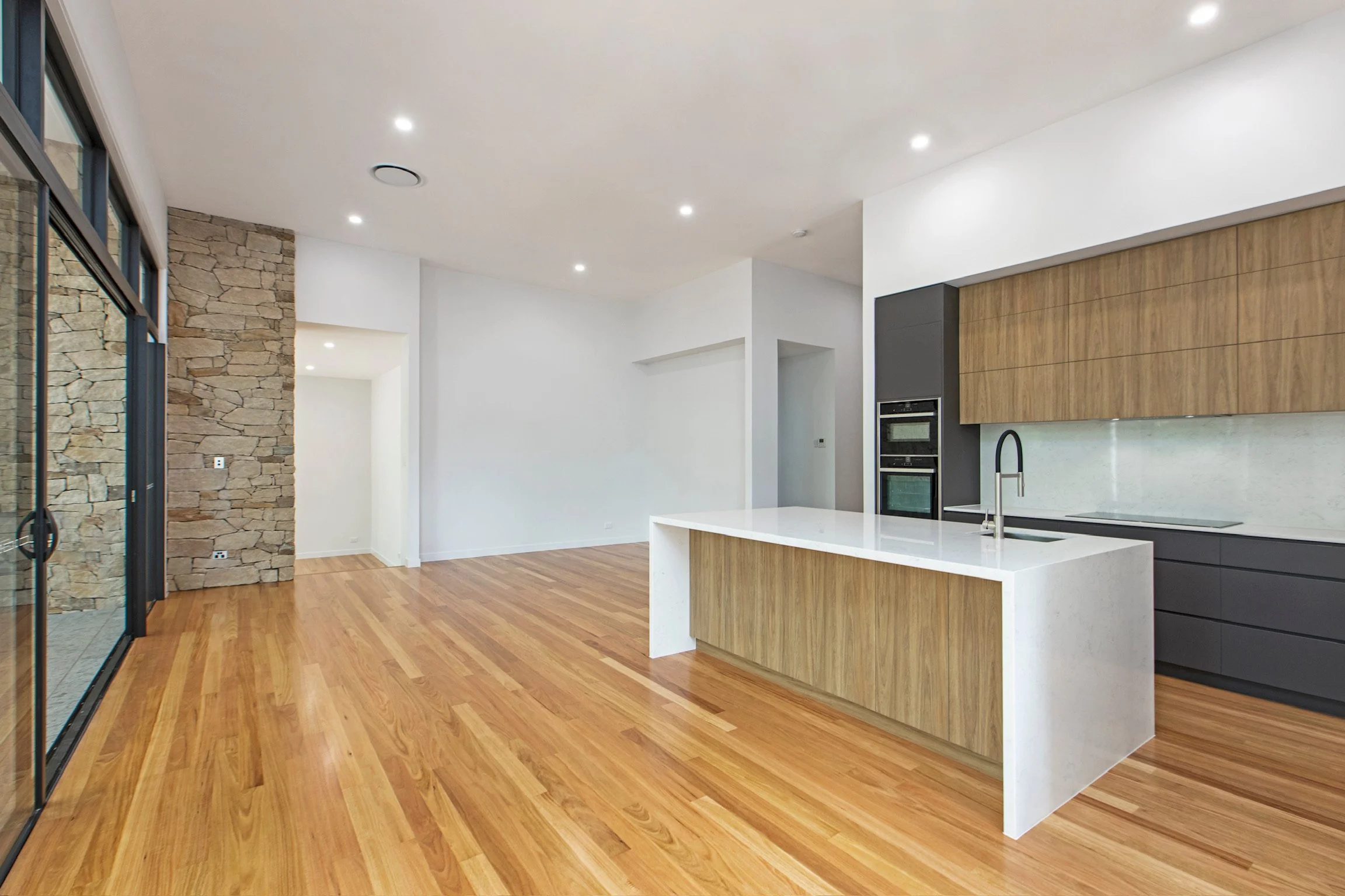 Modern open-concept kitchen and living space with hardwood floors, white walls, a stone accent wall, and built-in appliances, illuminated by recessed ceiling lights.