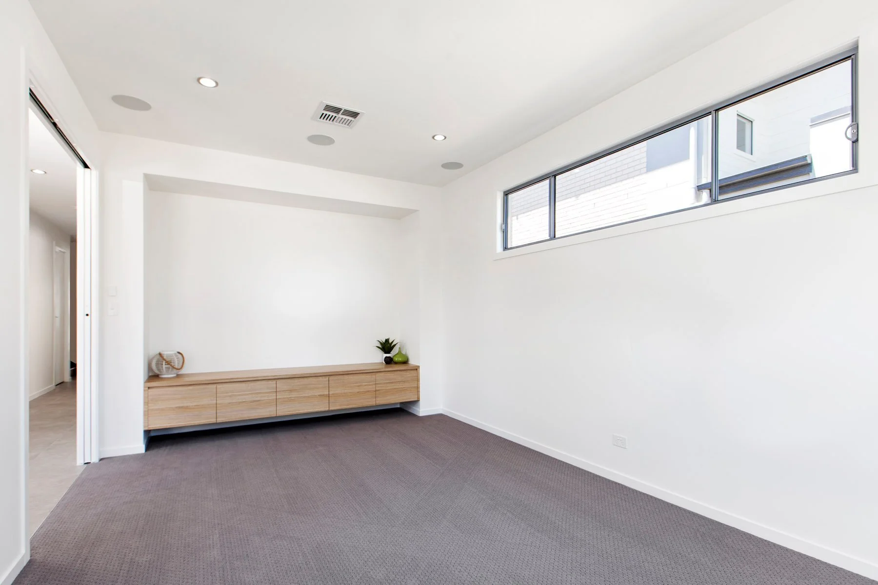 Empty modern room with white walls, a long horizontal window, short wood cabinet with a small plant and decorative objects, and a brown carpeted floor.