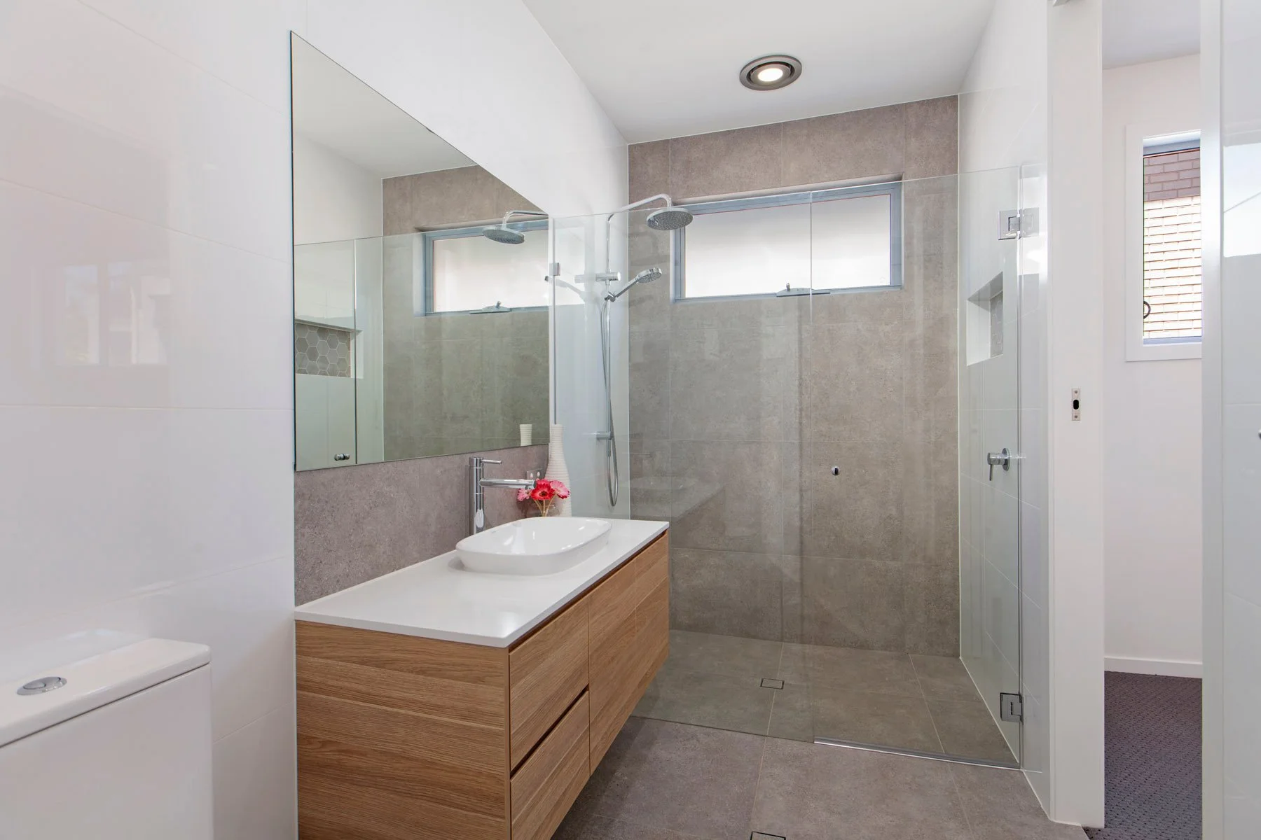 Modern bathroom with a large mirror, a white sink on a wooden vanity, a walk-in shower with glass walls, gray tile floors and walls, small windows, and decorative pink flowers on the vanity.