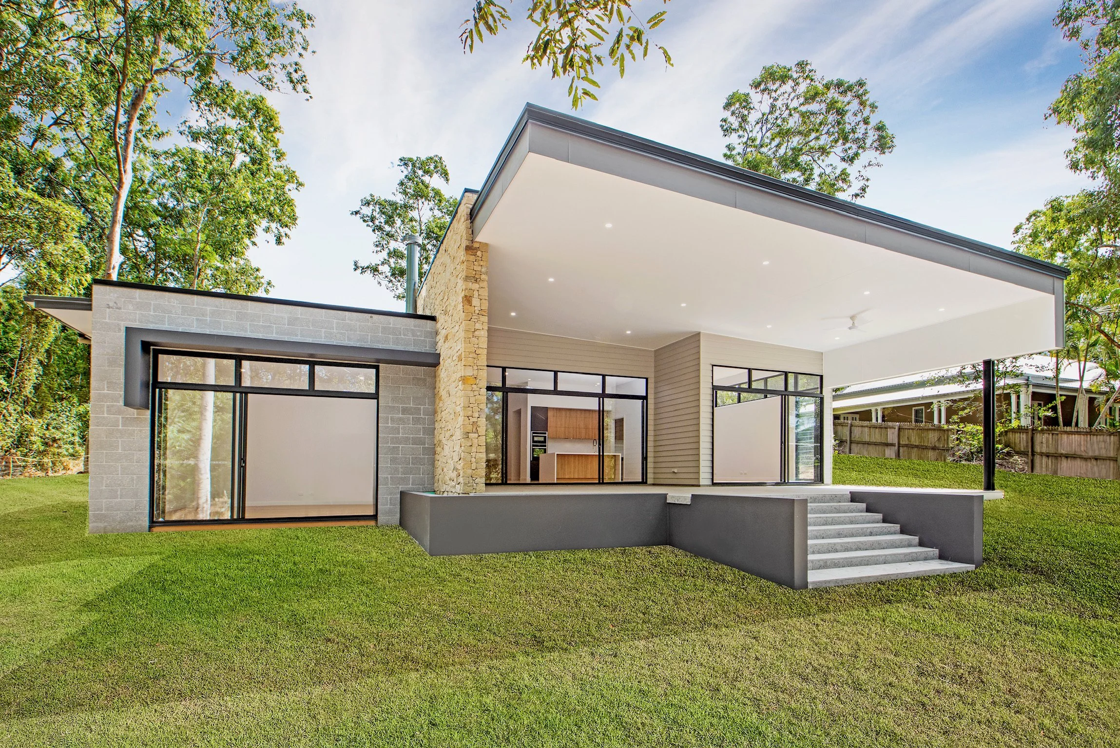 Modern house with large glass windows and door, stone accent wall, and a spacious covered patio with steps leading down to a green lawn, surrounded by tall trees under a partly cloudy sky.