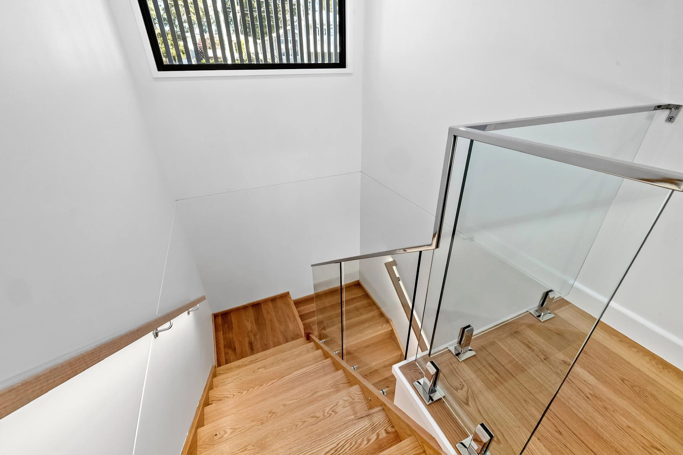 Interior view of a staircase with wooden steps, glass railing, and white walls, leading upward with a window at the top showing trees outside.