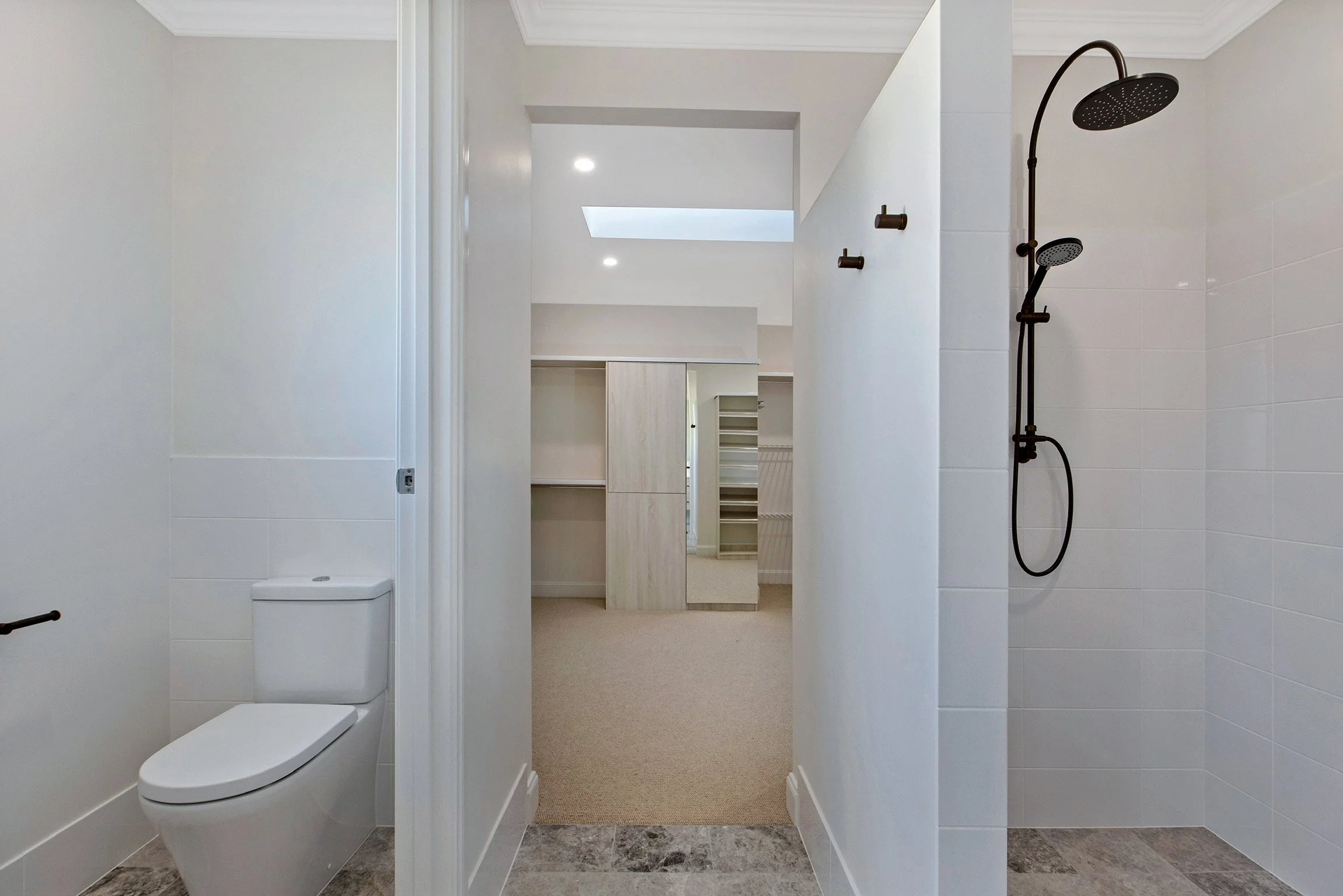 Modern bathroom with white tiled walls, a wall-mounted toilet, a walk-in shower with a black rain showerhead, and built-in shelves in the background.