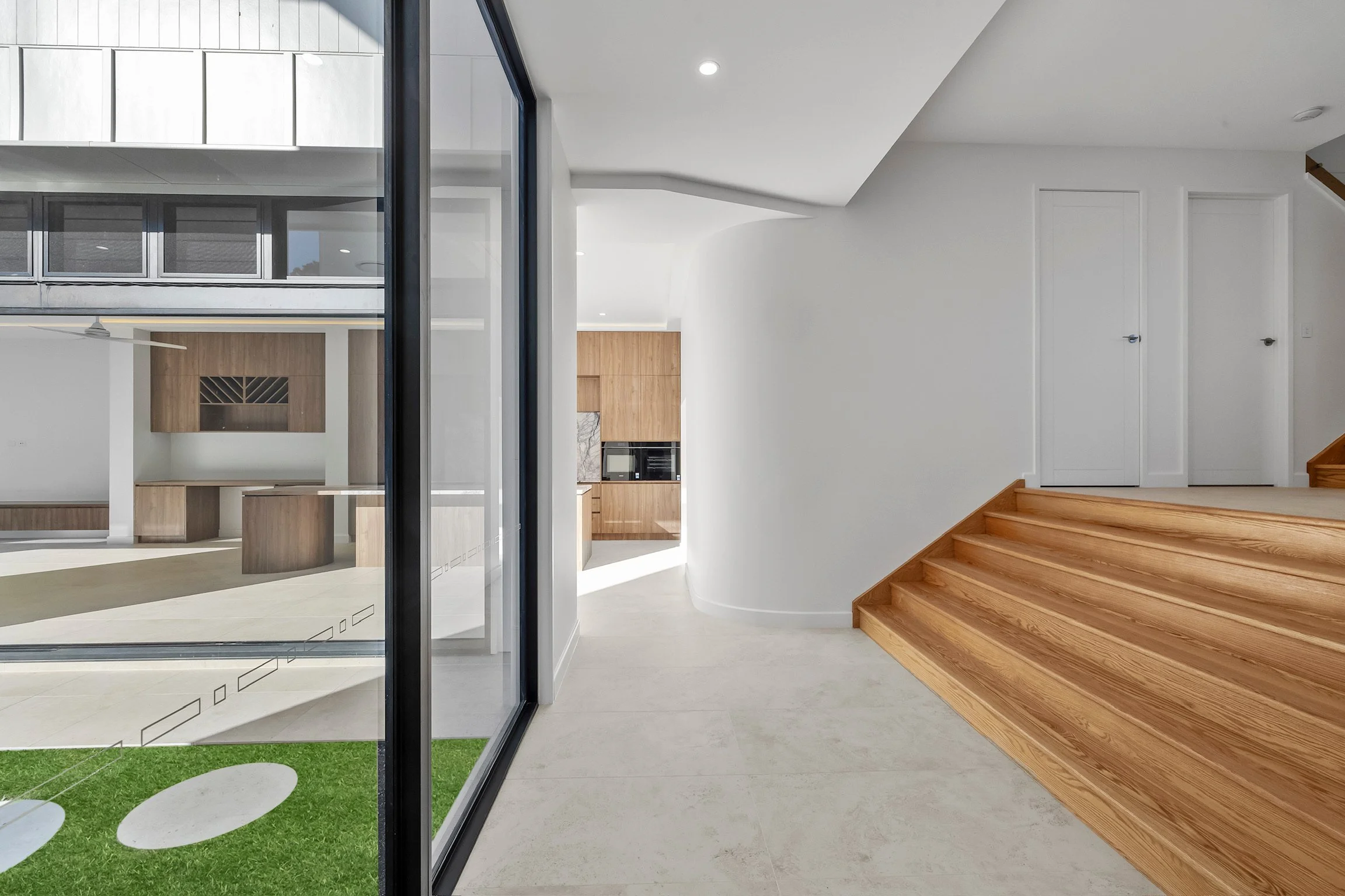 Modern interior with wooden stairs, white walls, a curved wall, and glass sliding door leading outside to a grassy area with stepping stones.
