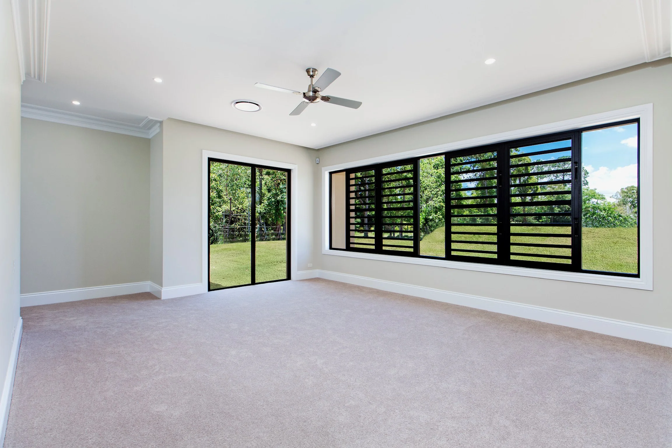 Empty room with beige carpet, light-colored walls, large black-framed windows, a sliding glass door, white crown molding, and a ceiling fan with lights.