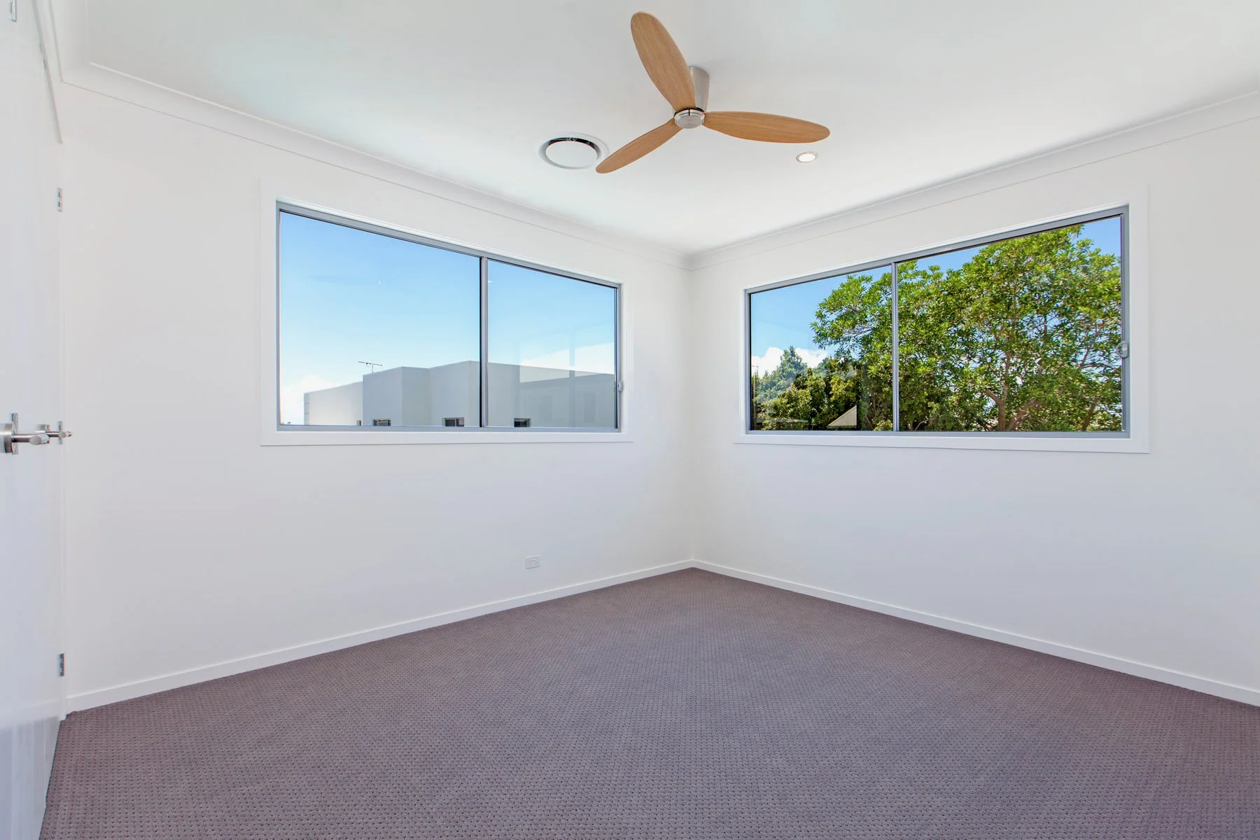 Empty room with white walls, grey carpet, and two large windows showing trees and a building outside. Ceiling has a wooden fan and recessed lighting.