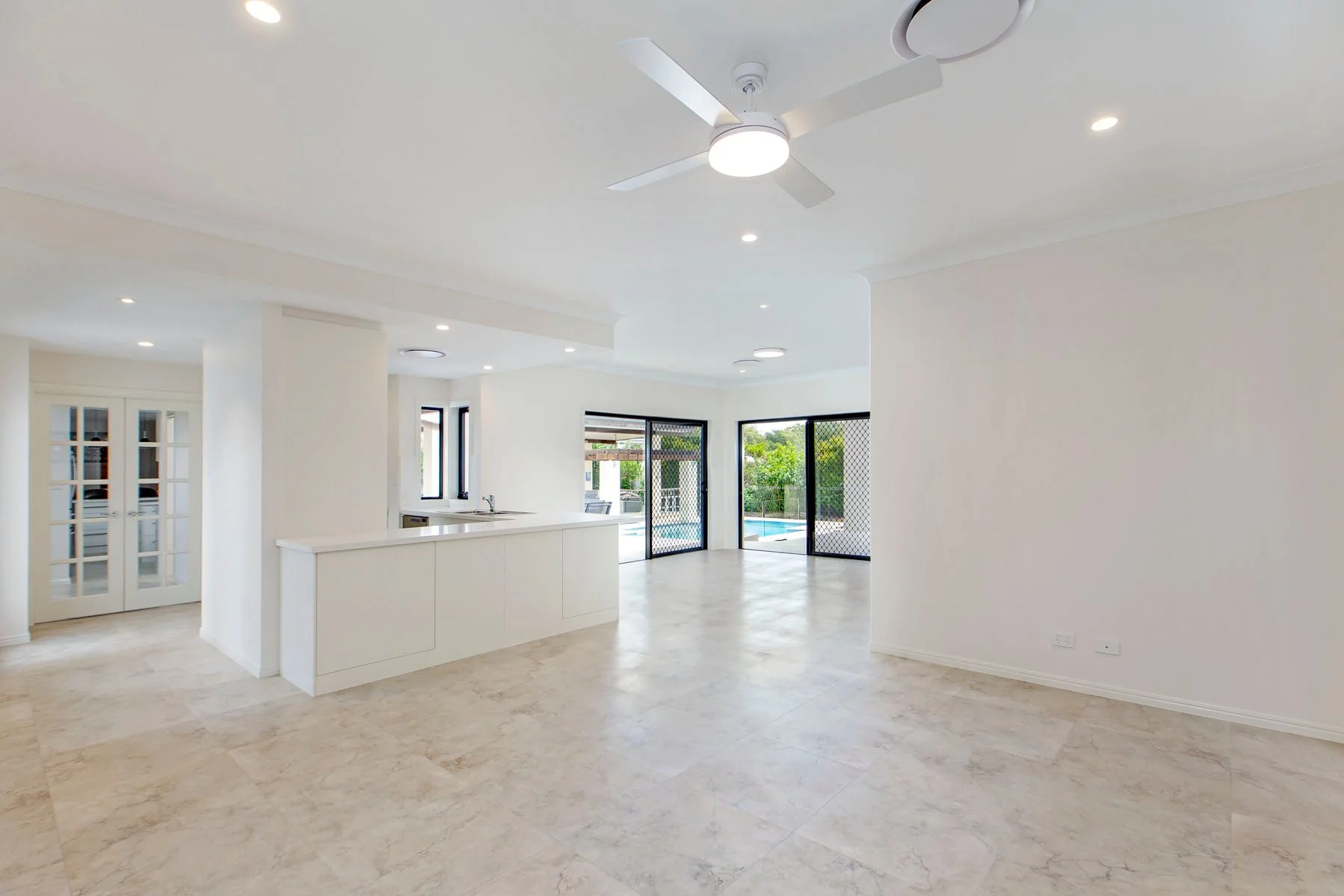 Empty living area with beige marble floors, white walls, a ceiling fan with lights, and sliding glass doors leading to a backyard pool.