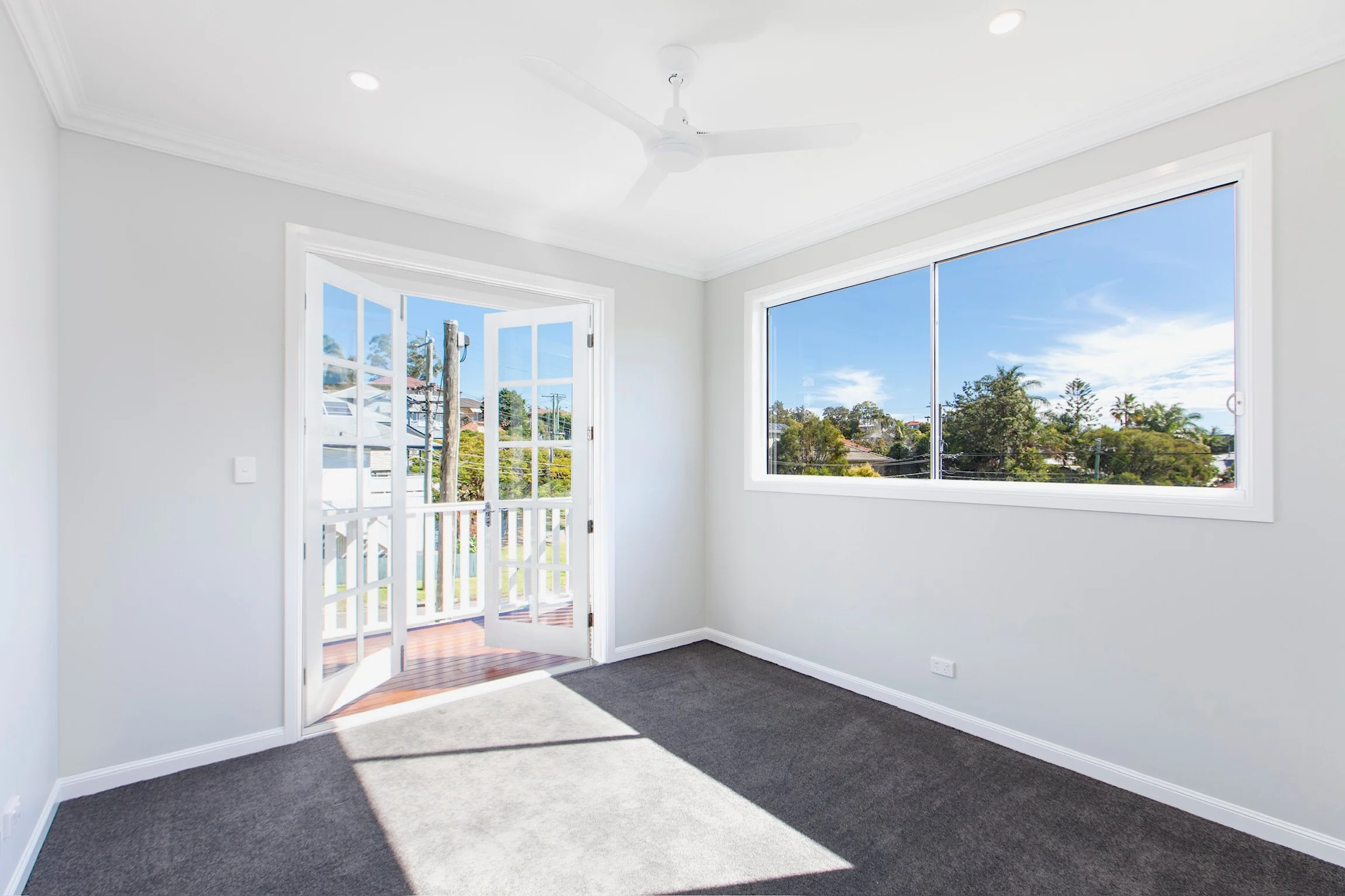 Empty room with large windows showing a sunny outdoor view, white walls, a ceiling fan, and gray carpeted flooring.