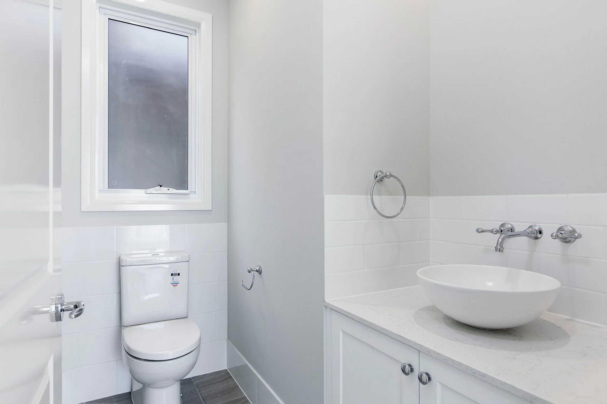 A modern bathroom with white walls and tiled lower walls. There is a window with frosted glass and a white frame, a toilet, and a countertop with a vessel sink. The bathroom has chrome fixtures, including a towel ring and faucet.