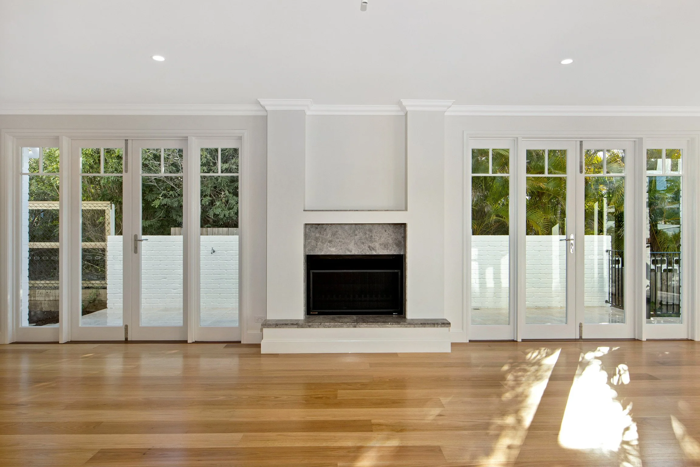 Empty living room with wooden floor, white walls, a fireplace, and large glass doors leading to the outdoor yard with trees.