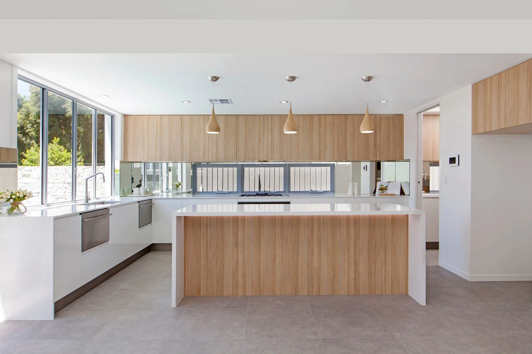 Modern kitchen with white countertops, wooden cabinets, large windows providing natural light, and pendant lights hanging from the ceiling.