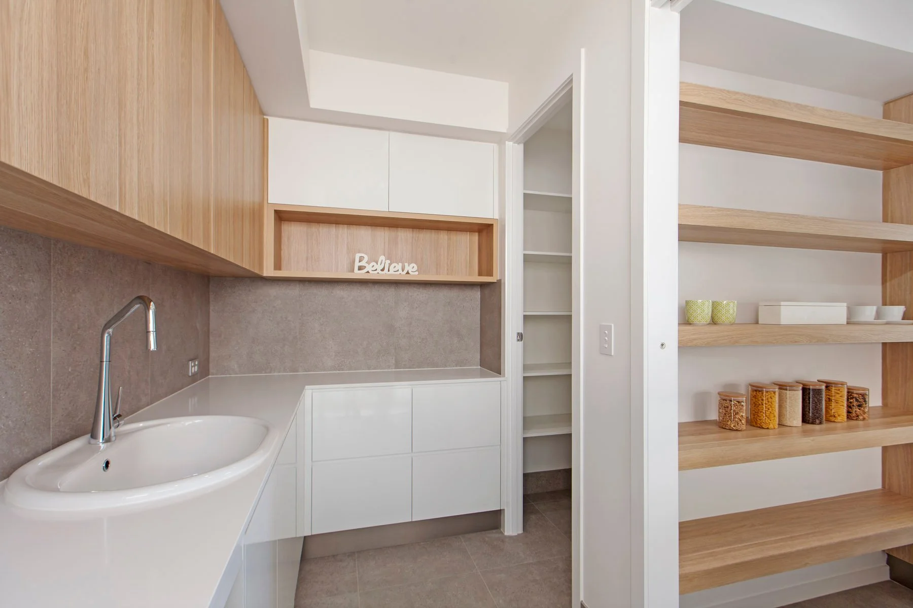 Modern kitchen with white countertops, wooden open shelves, and a pantry with glass jars of food.