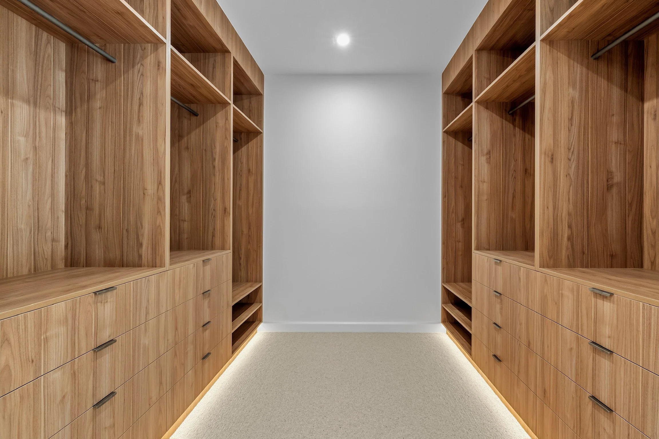 Empty walk-in closet with wooden shelves and drawers on both sides, beige carpet, white wall, and ceiling with a recessed light.