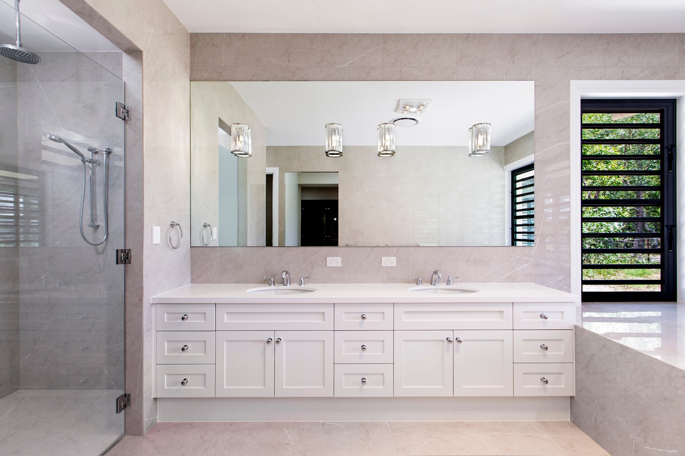 Modern bathroom with double vanity, large mirror, beige marble walls and flooring, wall-mounted lights, and a glass-enclosed shower with a rainfall showerhead.