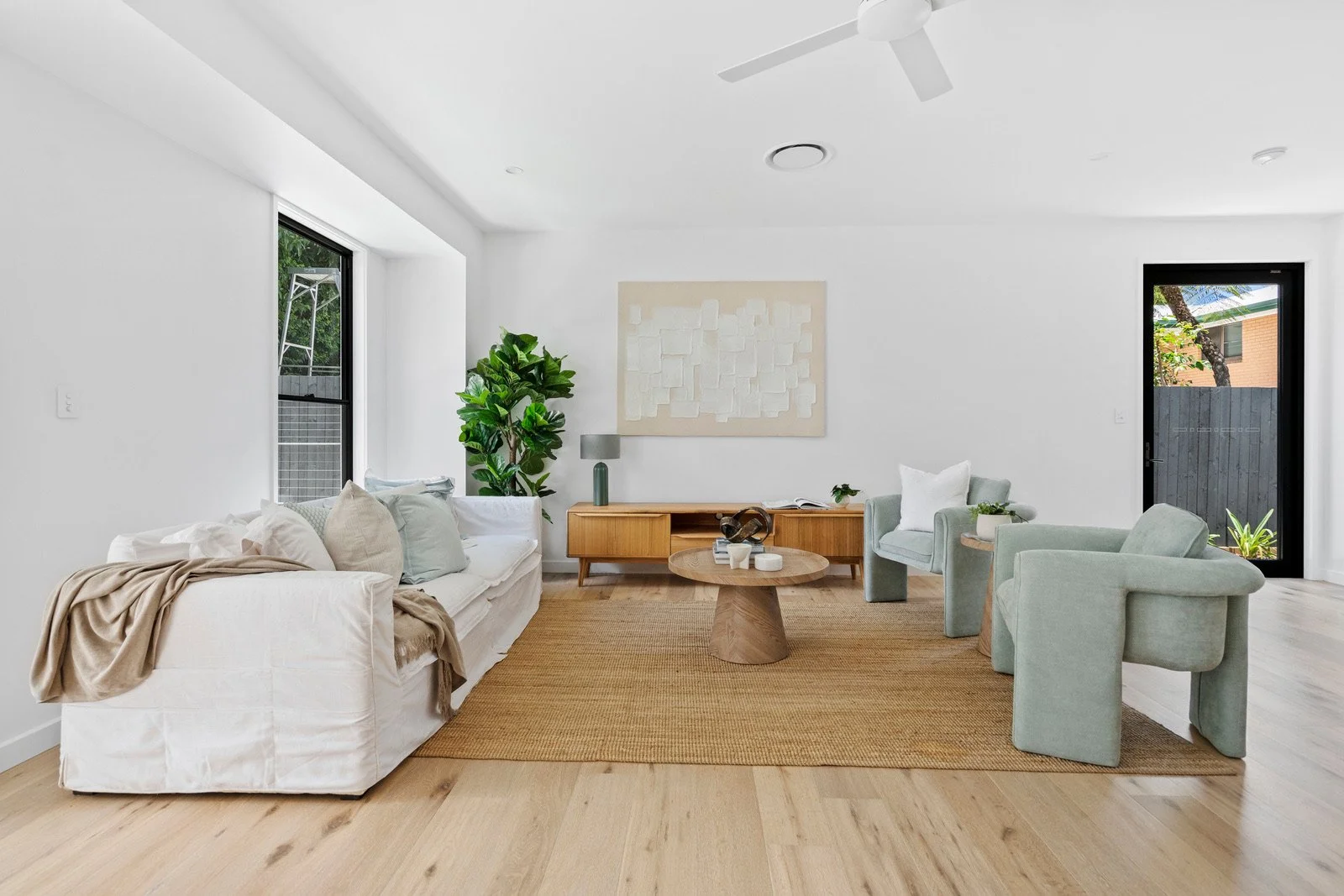 Modern living room with white walls and light wood flooring, containing a white sofa with beige and blue pillows, a beige blanket, two light green armchairs with pillows, a wooden coffee table, a wooden sideboard, a green potted plant, artwork on the