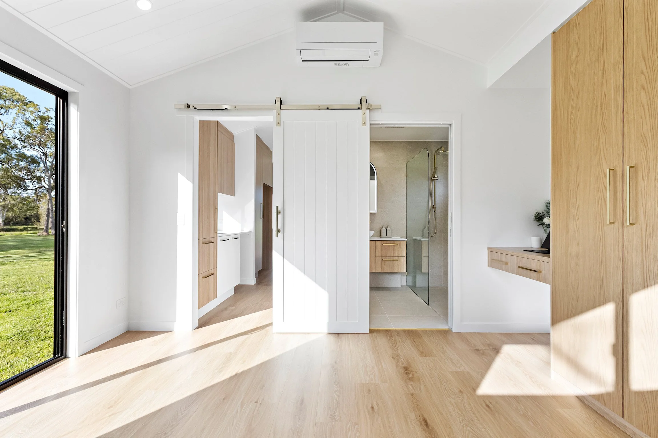 Bright, modern interior with light wooden floors, white walls, and a sliding barn door. Visible bathroom with a glass shower enclosure, and potted plant decor.