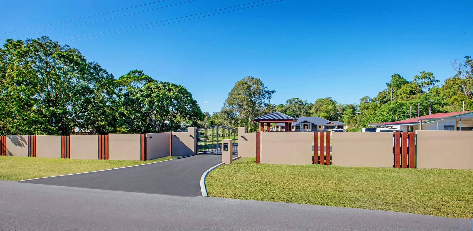 A gated residential community with a curved asphalt road, a decorative wall, green grass, leafy trees, and houses with colorful roofs.