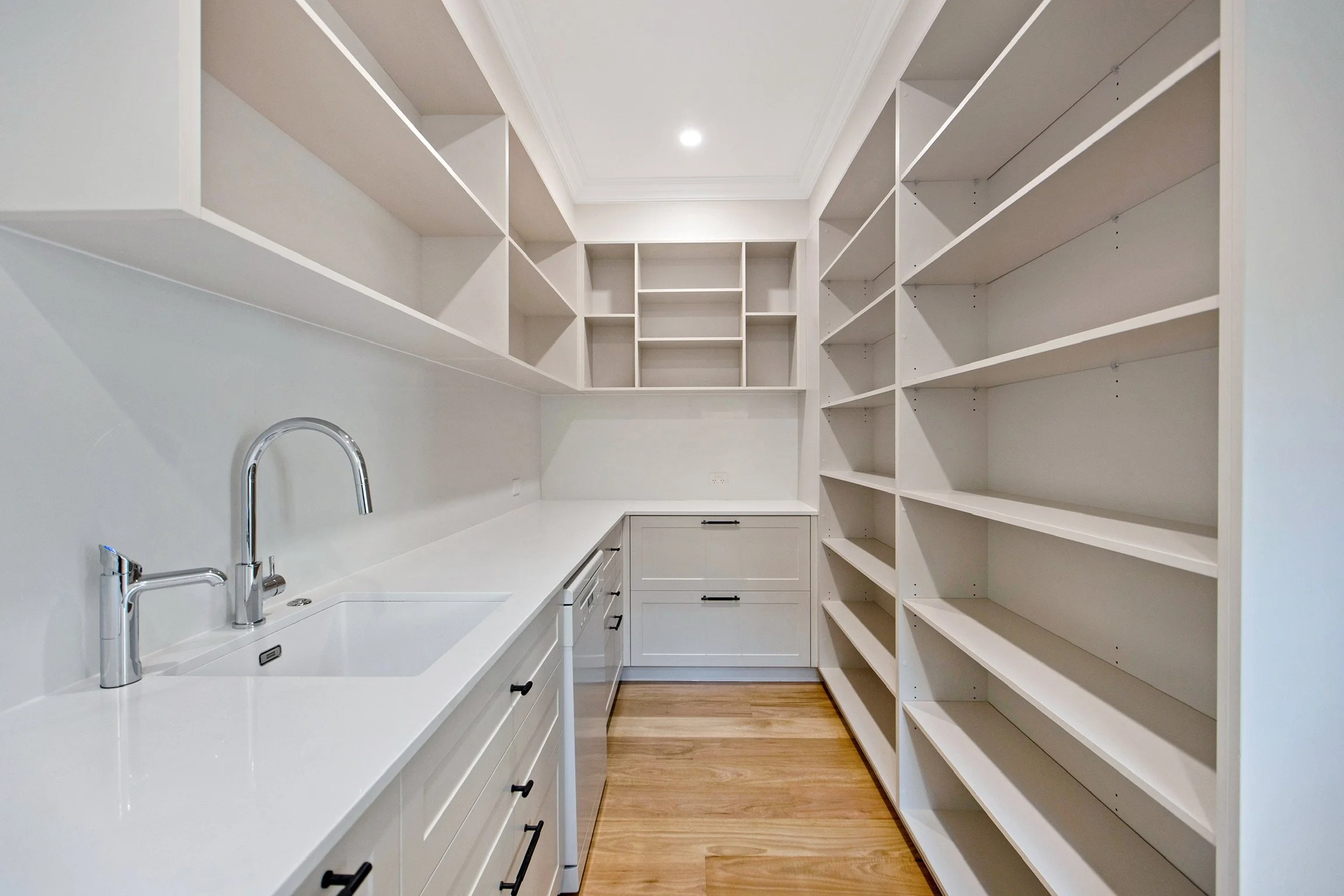 A modern, minimalist laundry or pantry room with white cabinets and open shelves on both sides, a white countertop with a sink and chrome faucet, wood flooring, and a ceiling light.