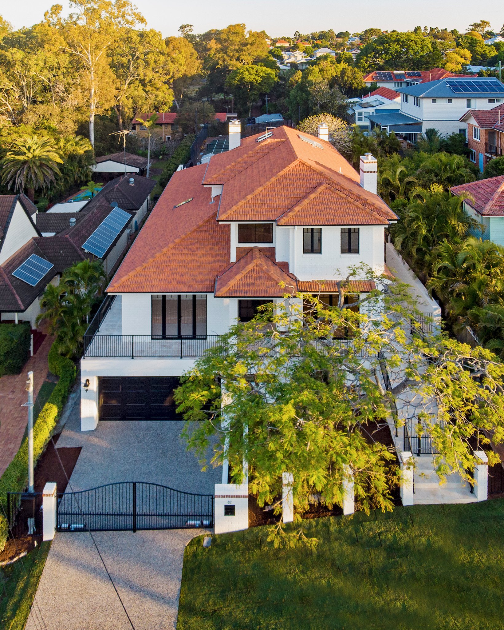 Aerial view of a modern house with white walls and a red tiled roof, surrounded by a gated yard, trees, and neighboring houses in a suburban neighborhood.
