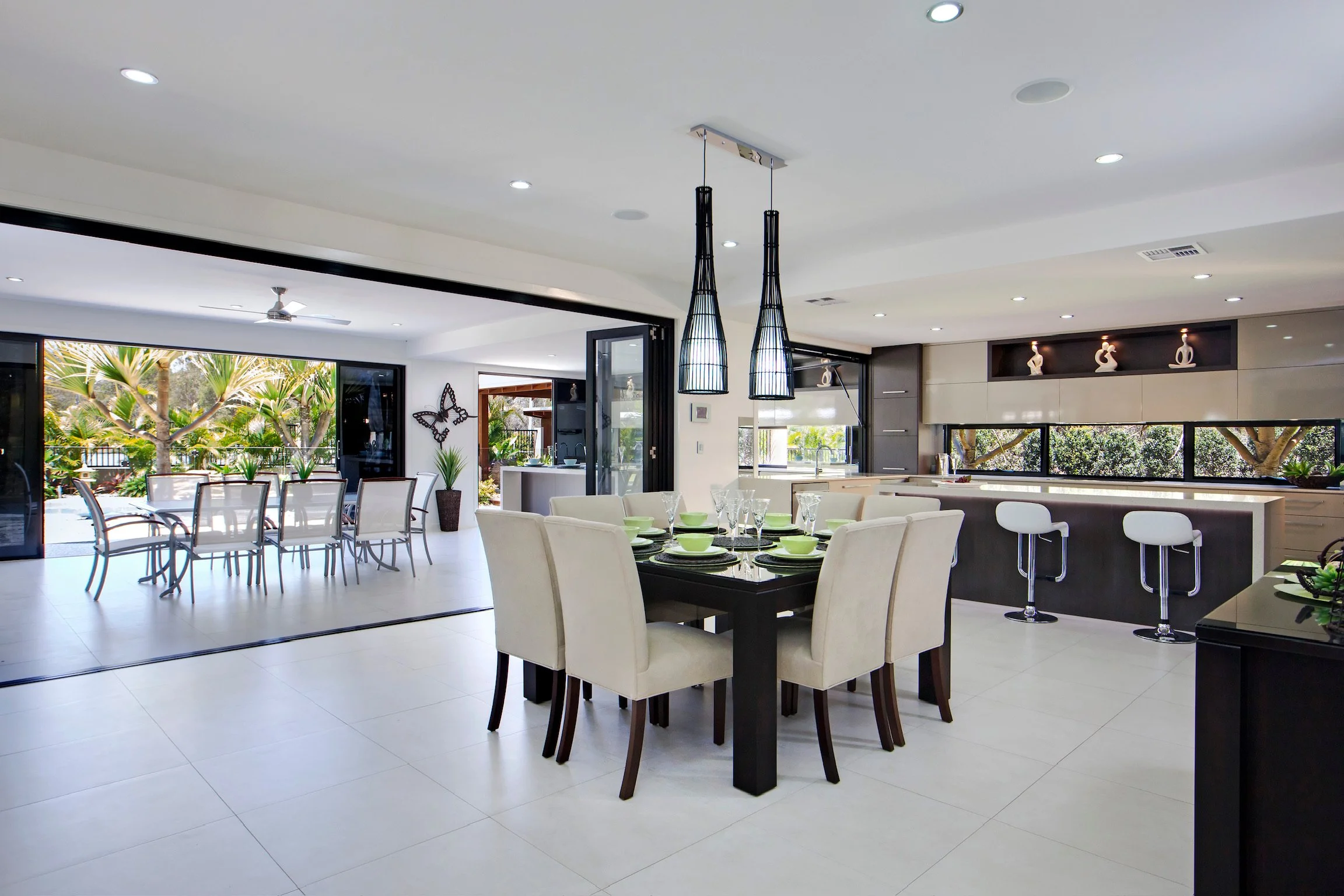 Modern open-concept kitchen and dining area with large windows, greenery outside, pendant lighting, and a table set with green and white dishware.