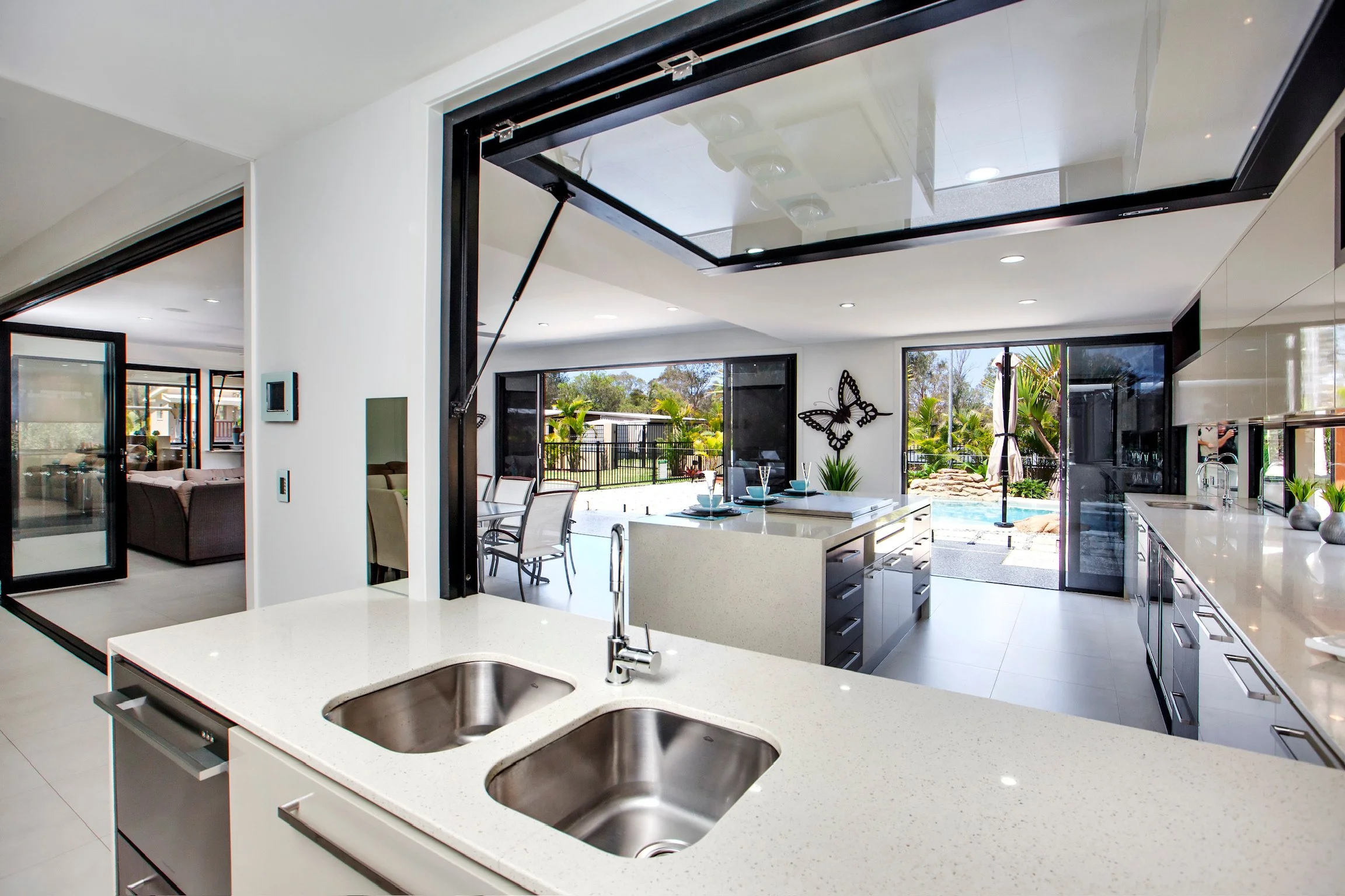 Modern kitchen with white countertops, stainless steel sinks, and white cabinetry. Large sliding glass doors lead to a backyard with a pool and outdoor seating area. Decor includes a black butterfly wall art piece.