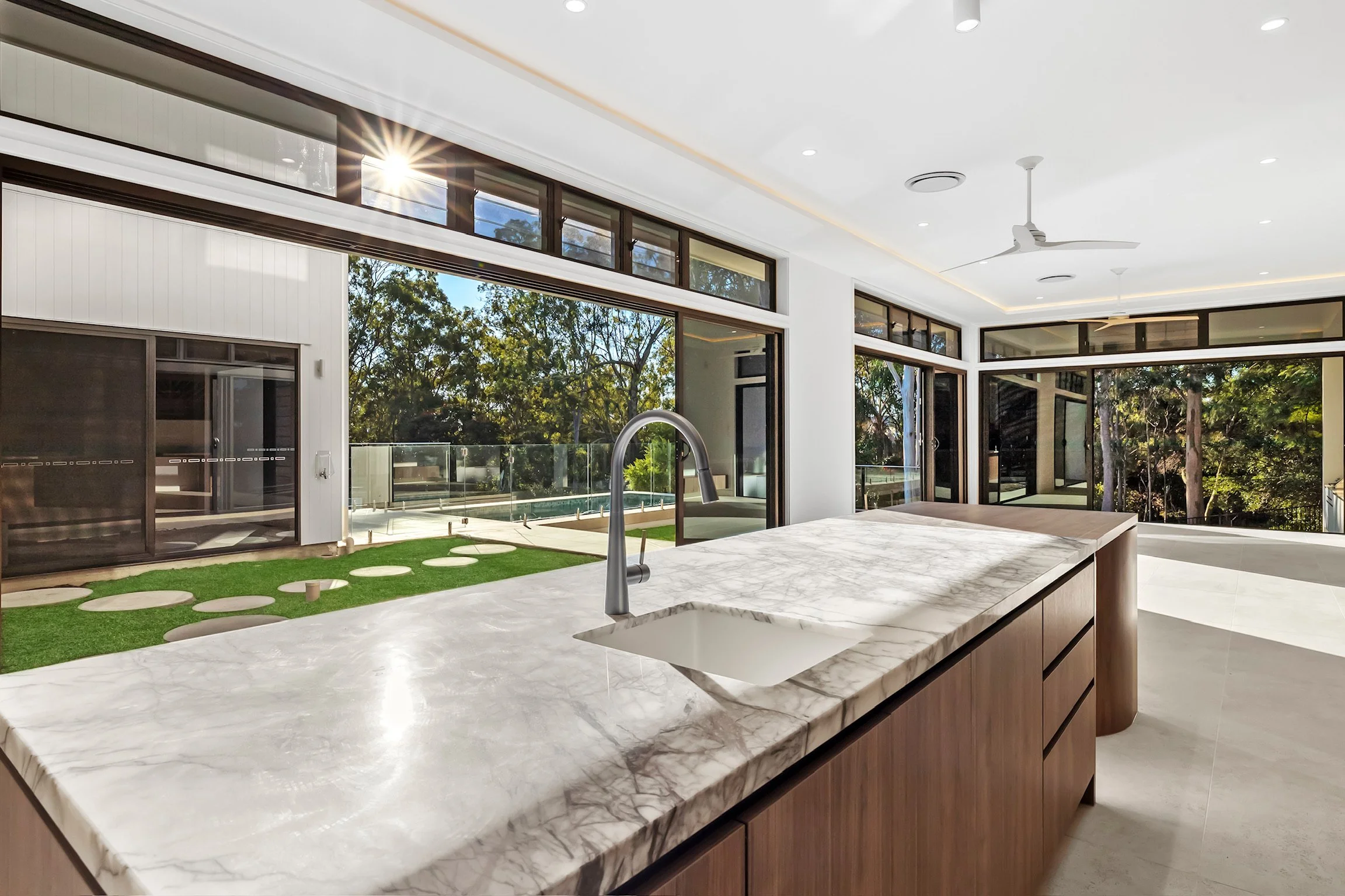 Modern kitchen with white marble island, wooden cabinets, large sliding glass doors, and a view of a backyard with trees and a deck.