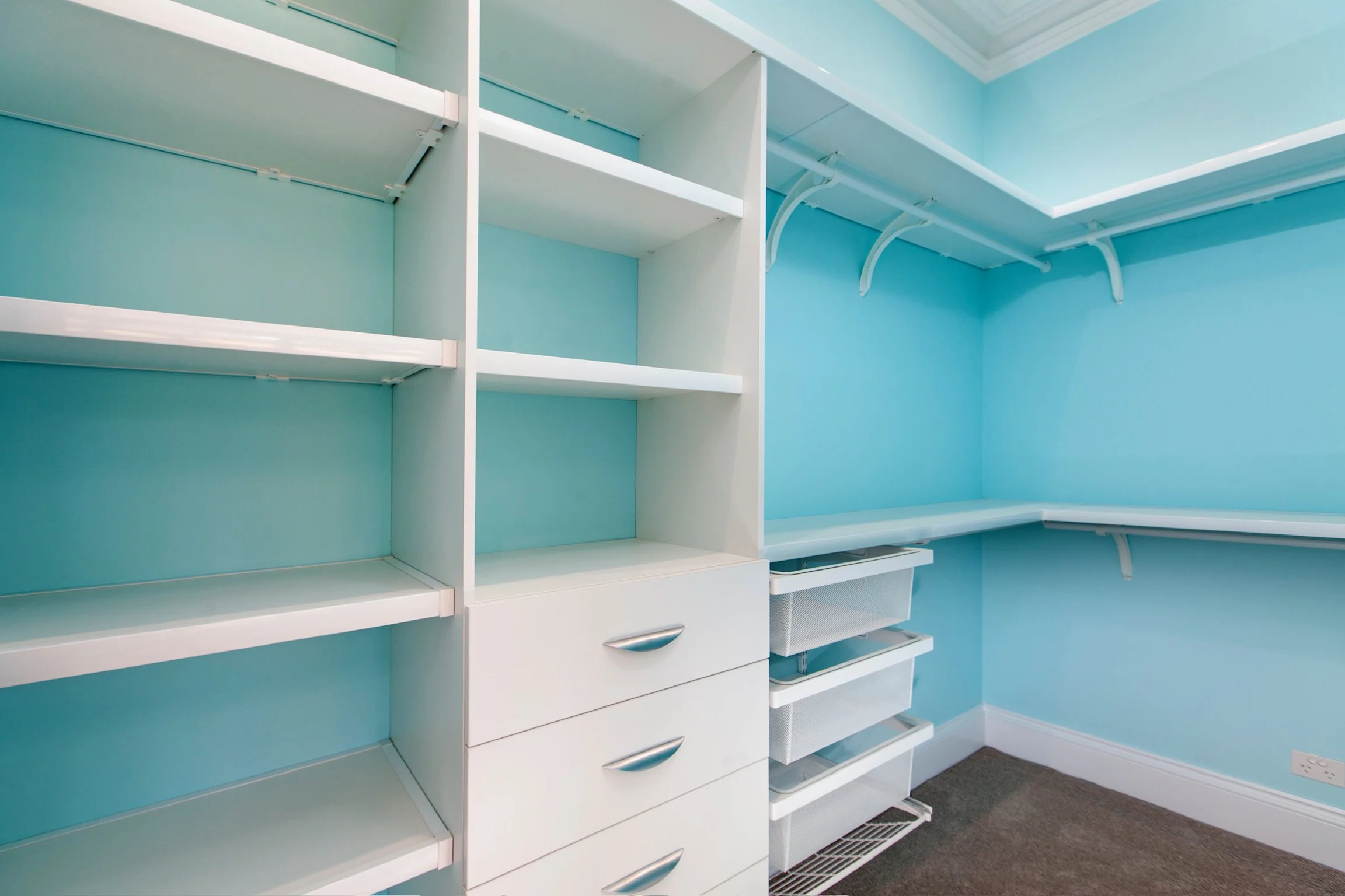 Empty closet with white shelving units and drawers, set against light blue walls with a vent on the dark gray carpeted floor.