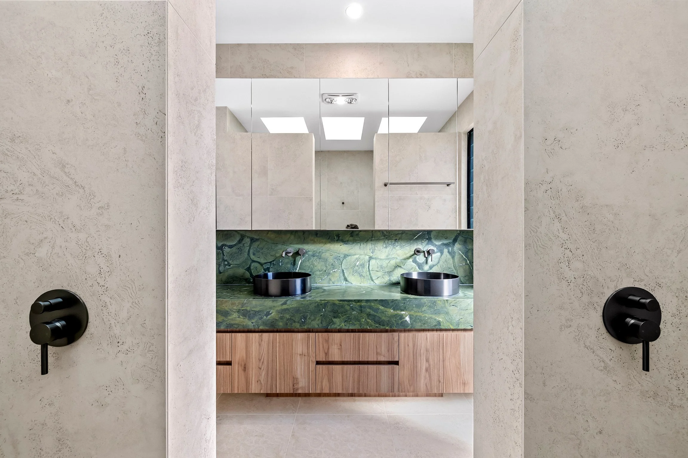 Modern bathroom with double sink vanity, green marble countertop, wooden cabinet below, large mirror, wall-mounted black faucets, and textured beige walls.