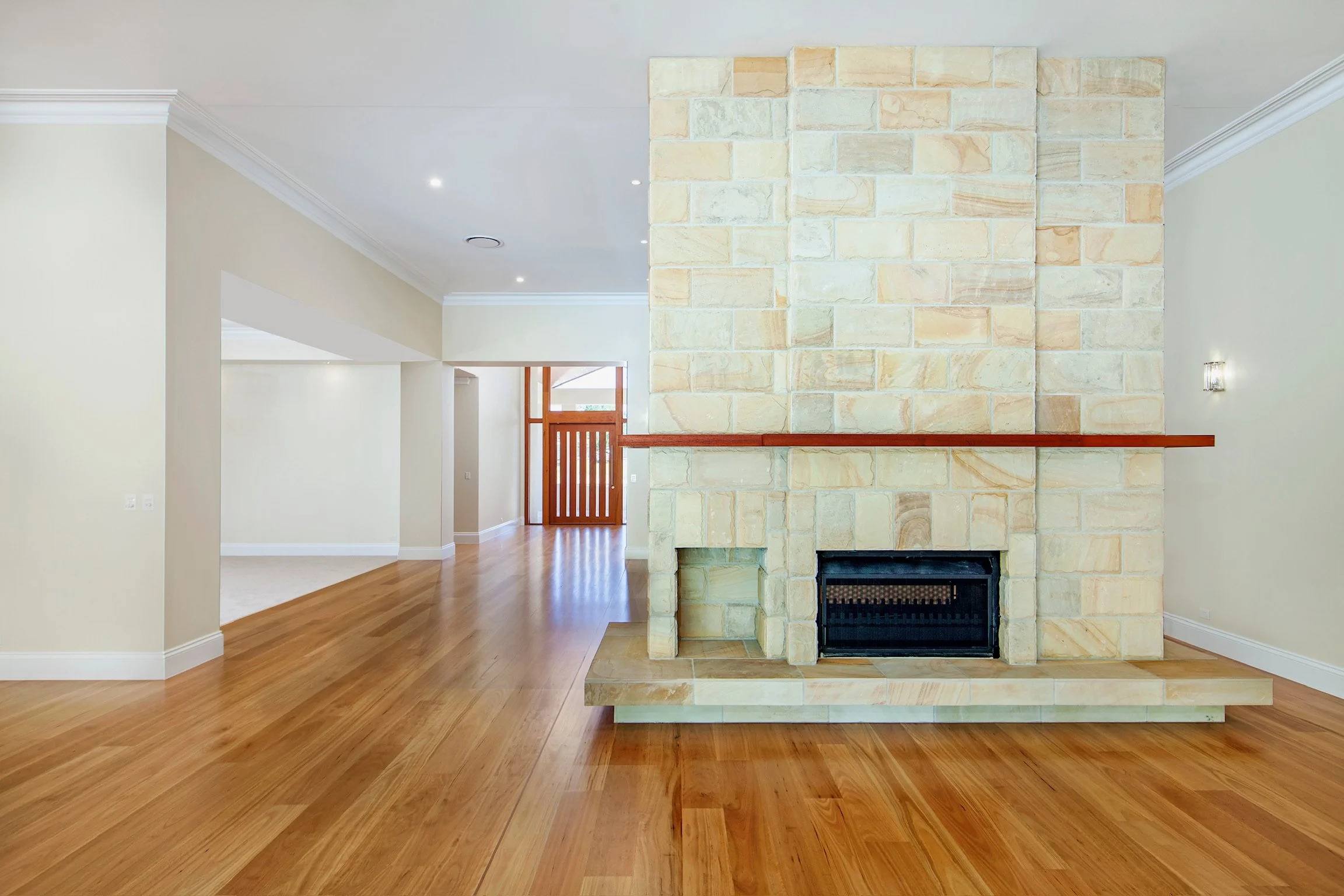 Interior of a house featuring a large stone fireplace with a wooden mantel, hardwood floors, and off-white walls. In the background, there are visible doorways and a wooden front door with glass panels.