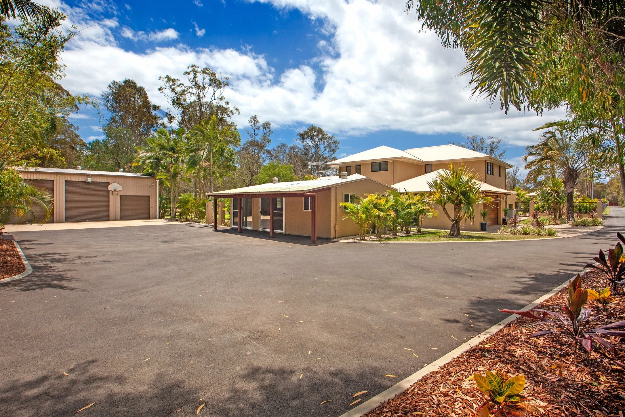 A residential parking lot with a beige building and a detached garage, surrounded by tropical trees and plants under a partly cloudy sky.