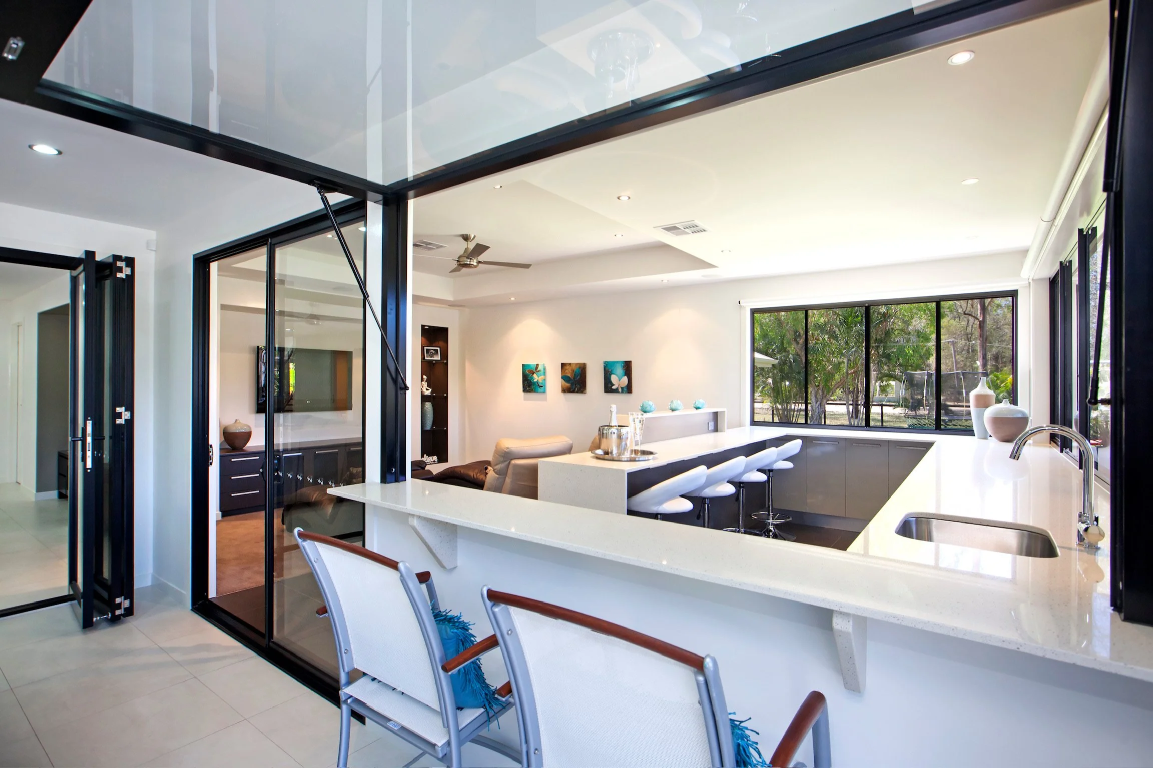 Modern kitchen and living room with large windows, white countertops, bar seating, and artwork on the walls.