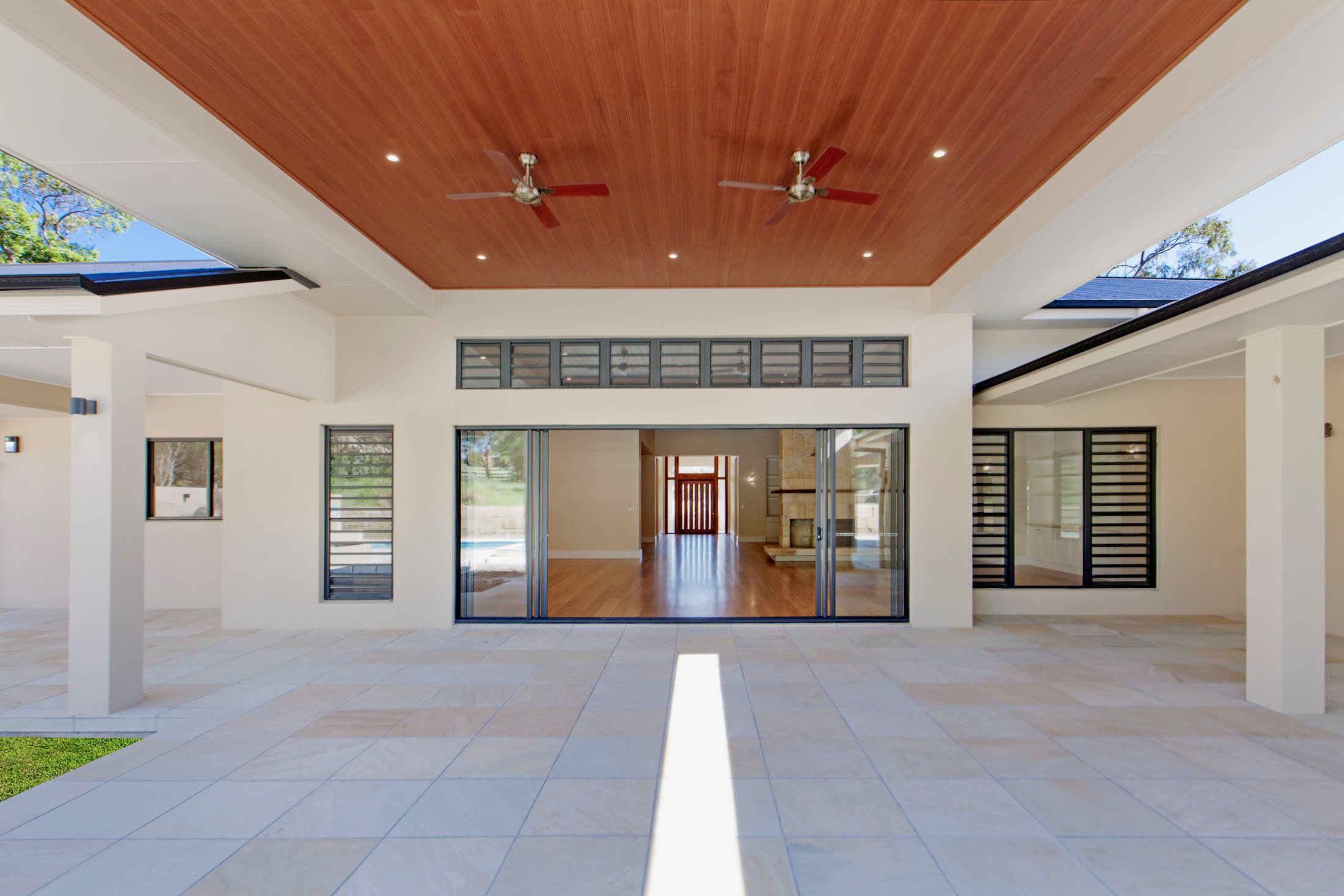 Modern open-concept living area with large sliding glass doors, tiled floor, and a wood-paneled ceiling with ceiling fans and recessed lighting.