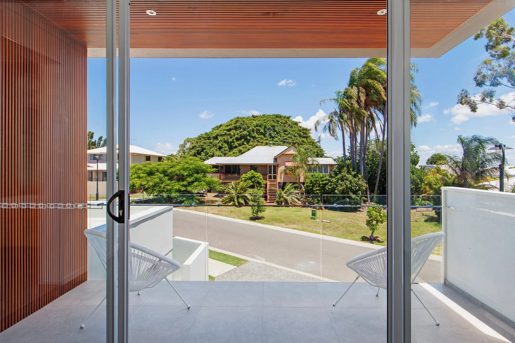 View from a balcony showing a suburban neighborhood with houses, tall trees, and a clear blue sky, framed by a sliding glass door and a wooden ceiling.