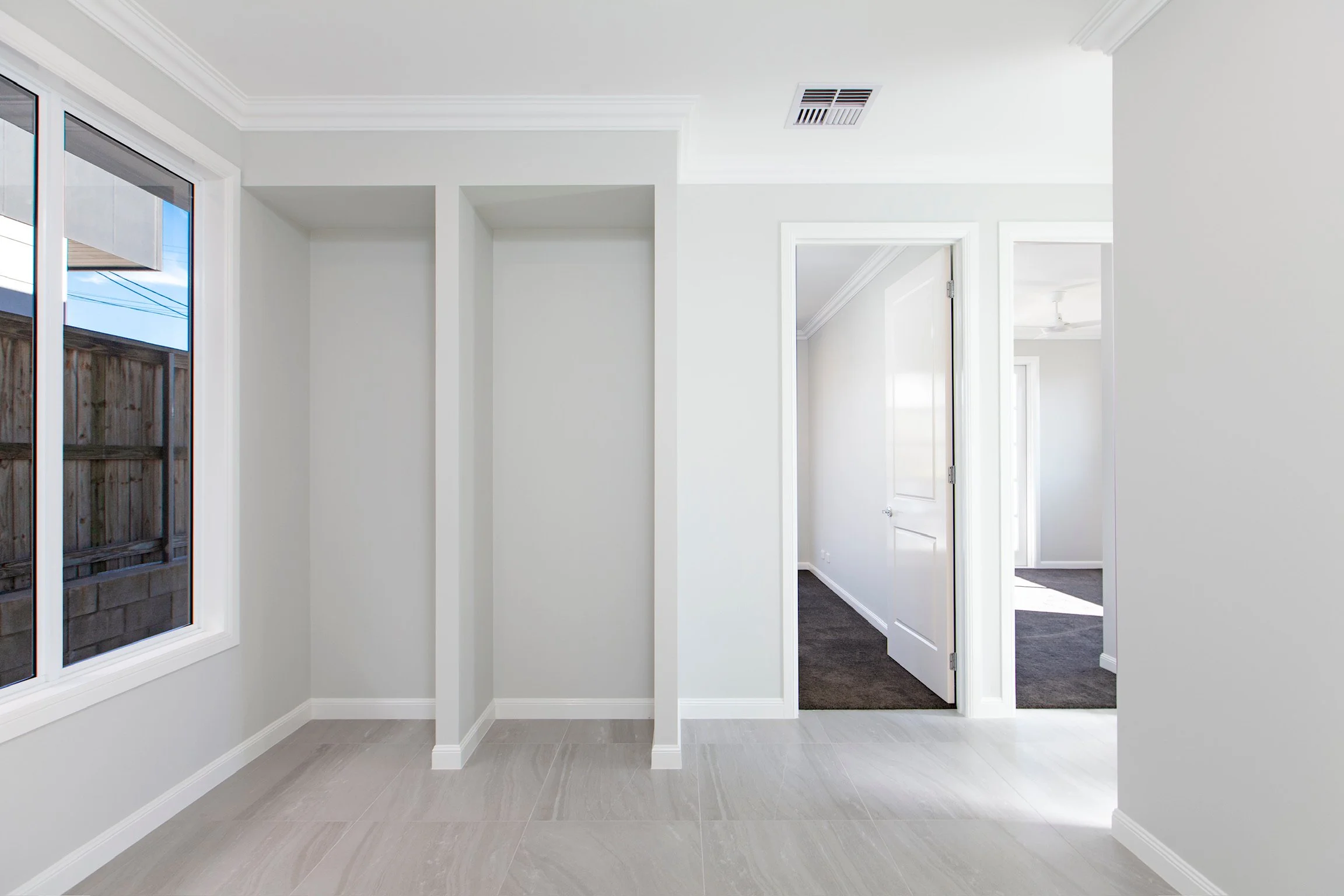 Interior view of a modern, bright house with white walls, a window showing a wooden fence outside, multiple doorways, crown molding, and light-colored flooring.