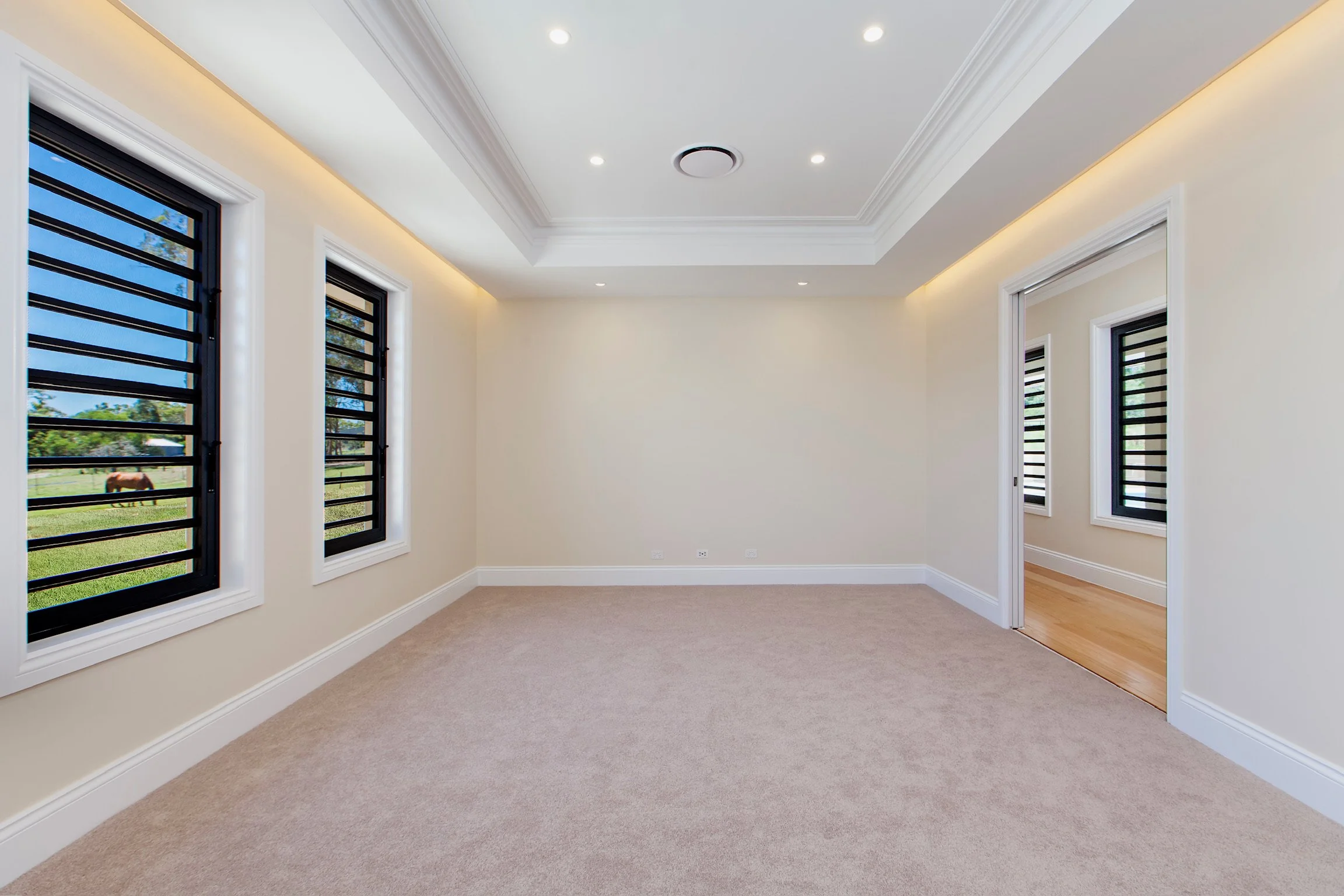 Empty bright room with large windows, beige walls, white baseboards, and a carpeted floor. There is a door opening to another room with wooden flooring.